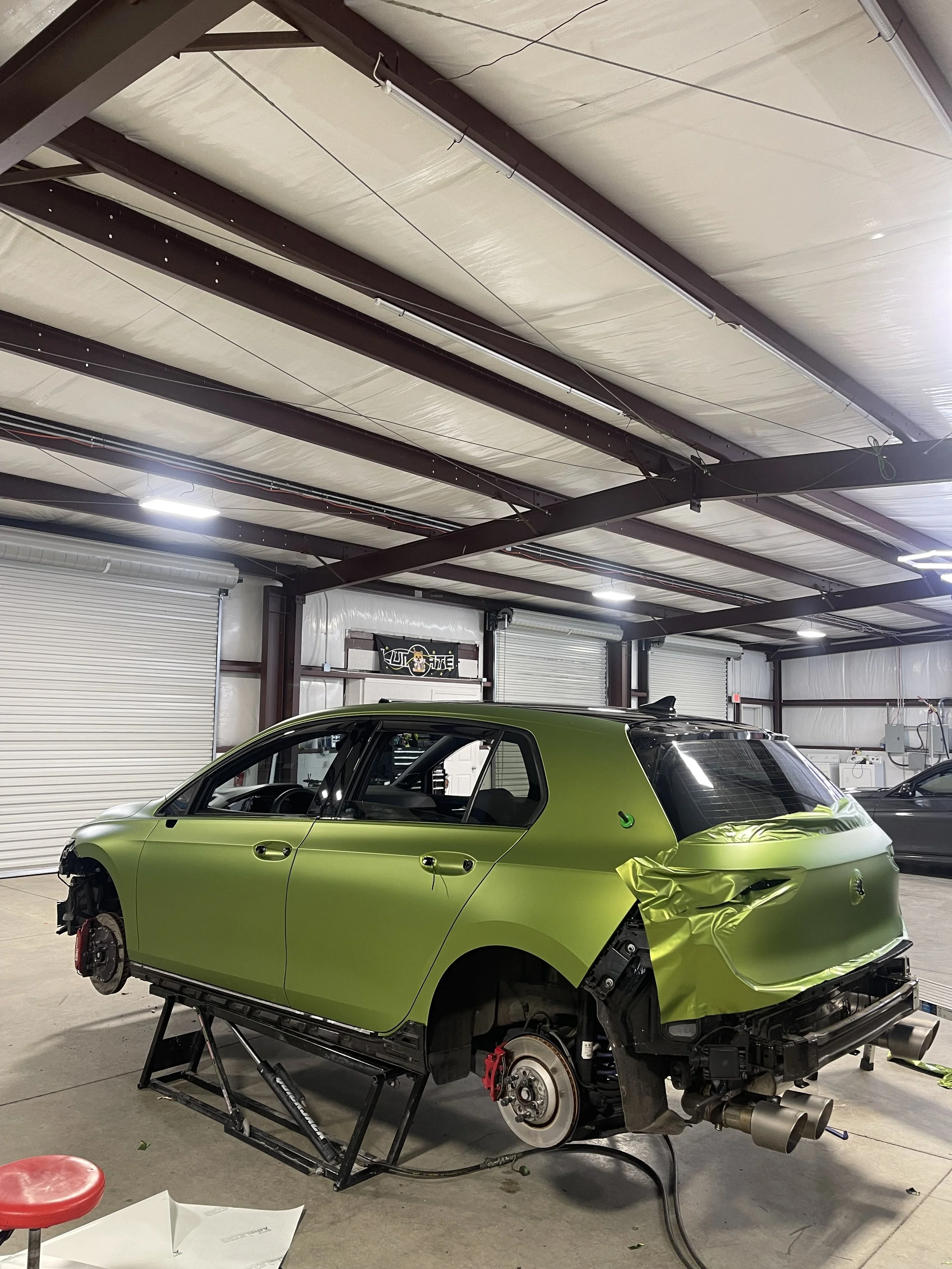 A green hatchback car in a garage undergoing repairs, missing wheels, with exposed brake parts and sans rear bumper, with tools and equipment nearby.