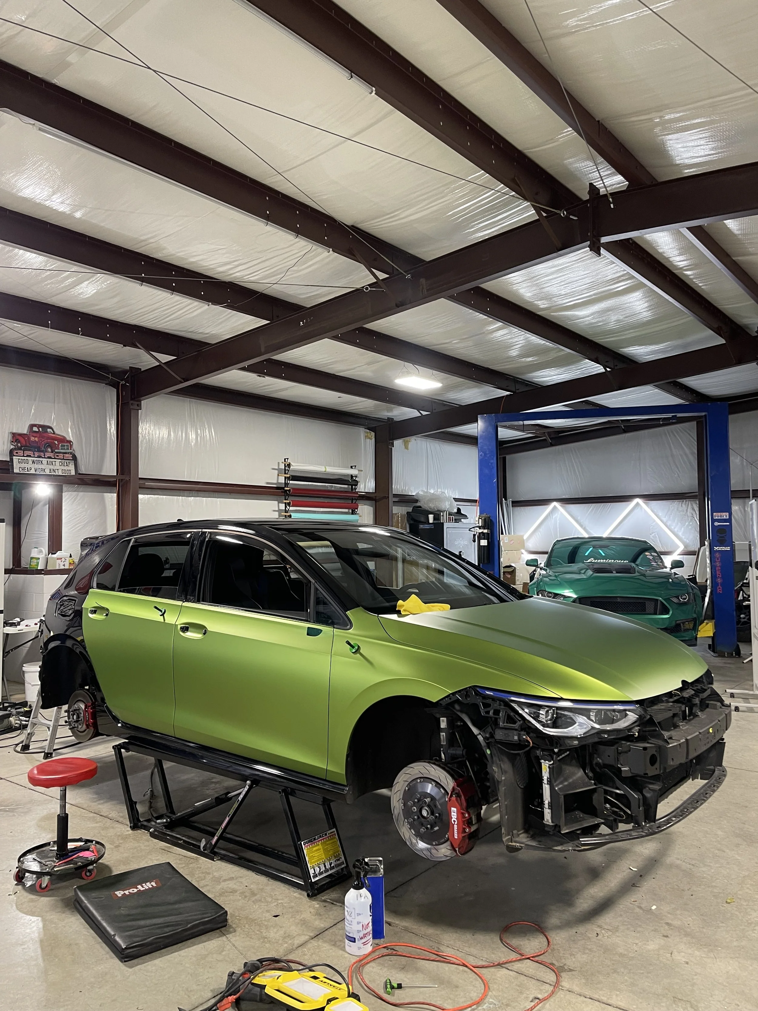 A car in the process of repair inside a garage, with the front wheels removed and the car supported on a lift. The car is lime green with the front bumper and other parts removed. There are various tools and equipment scattered around.