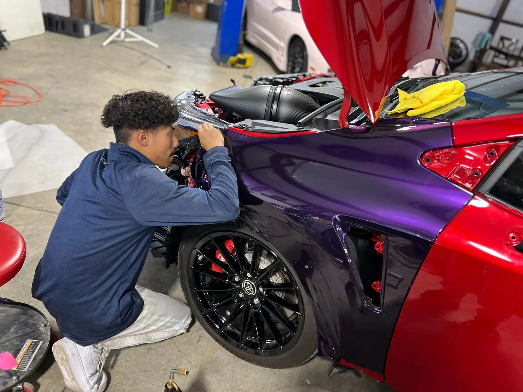 A young man working on the engine of a sports car with a red and purple exterior in a garage or workshop.