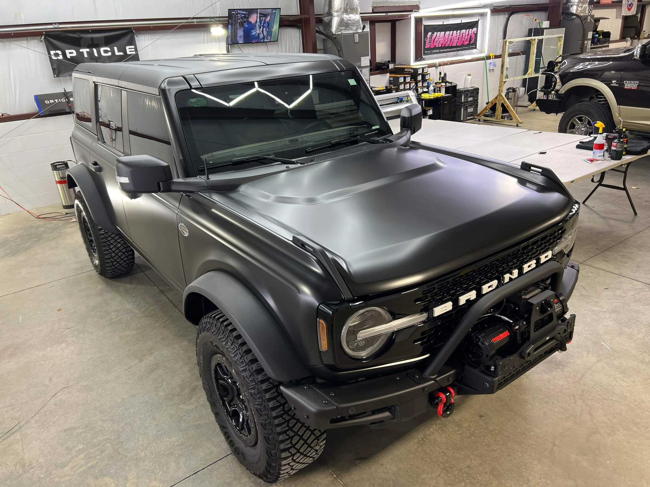 Black matte off-road vehicle with 'BRADONCO' on front grille, parked inside a workshop with tools and supplies on a table nearby.