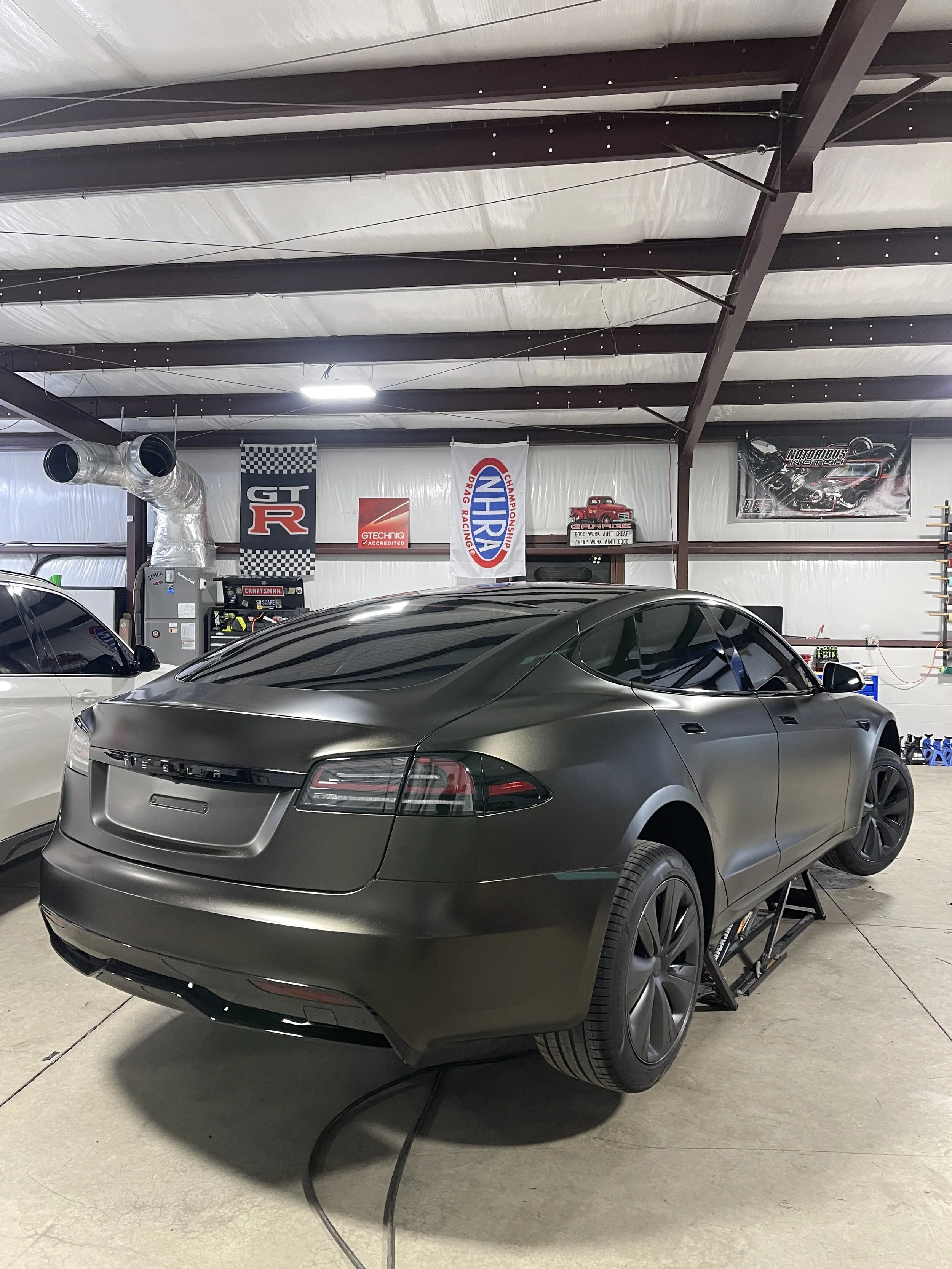 Black Tesla Model S in a garage, raised on a vehicle lift with a black matte finish, surrounded by racing banners, car parts, and tools.
