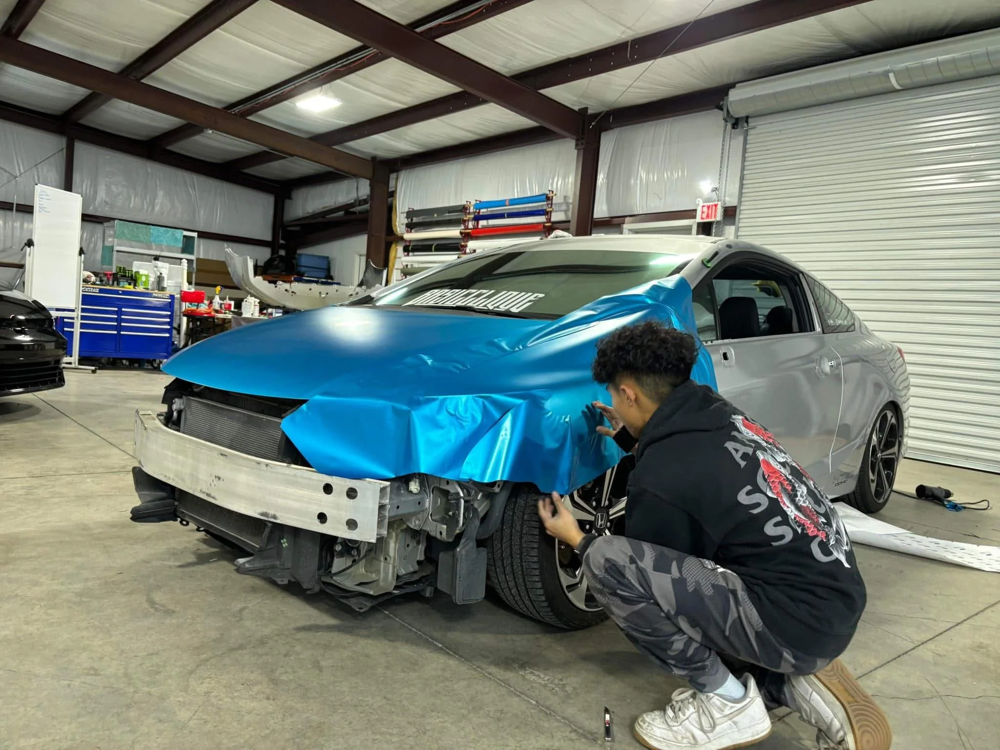 A person working on repairing a silver car with a blue protective cover in a garage or workshop.
