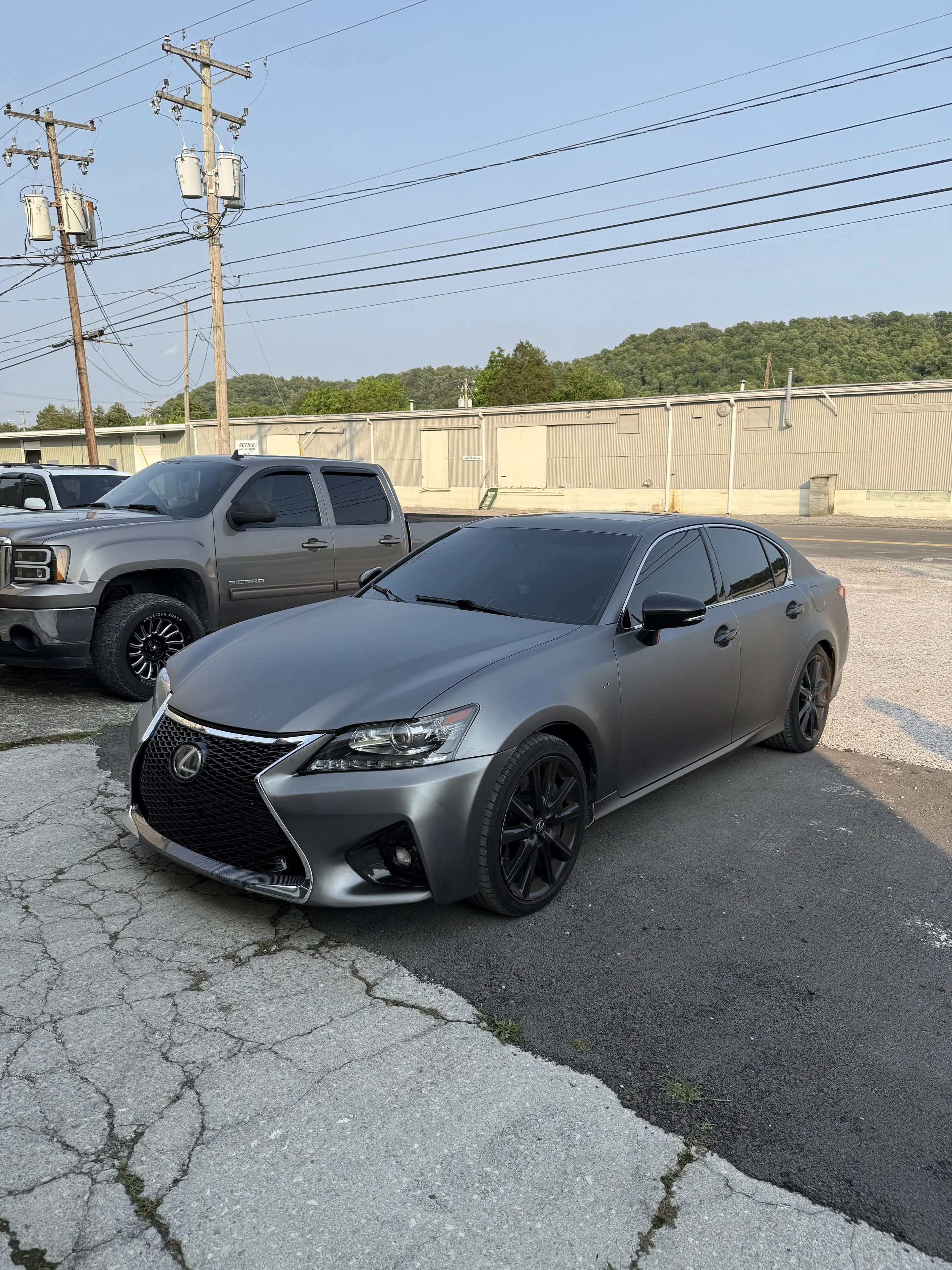 A gray Lexus sedan parked on cracked pavement beside a silver pickup truck with tinted windows, under power lines with green hills in the background.