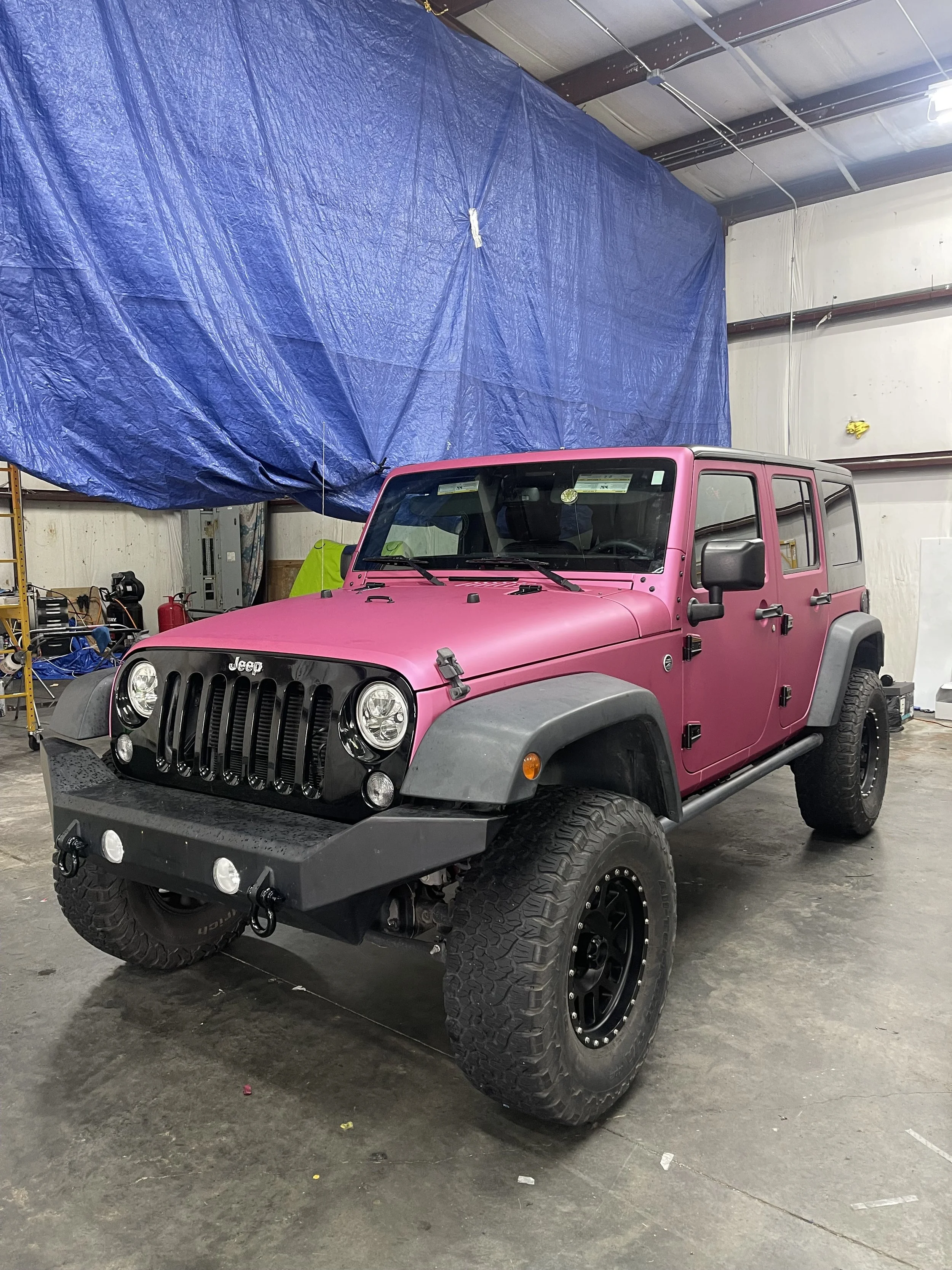 A pink Jeep Wrangler with black fender flares and large off-road tires parked inside a warehouse or garage.