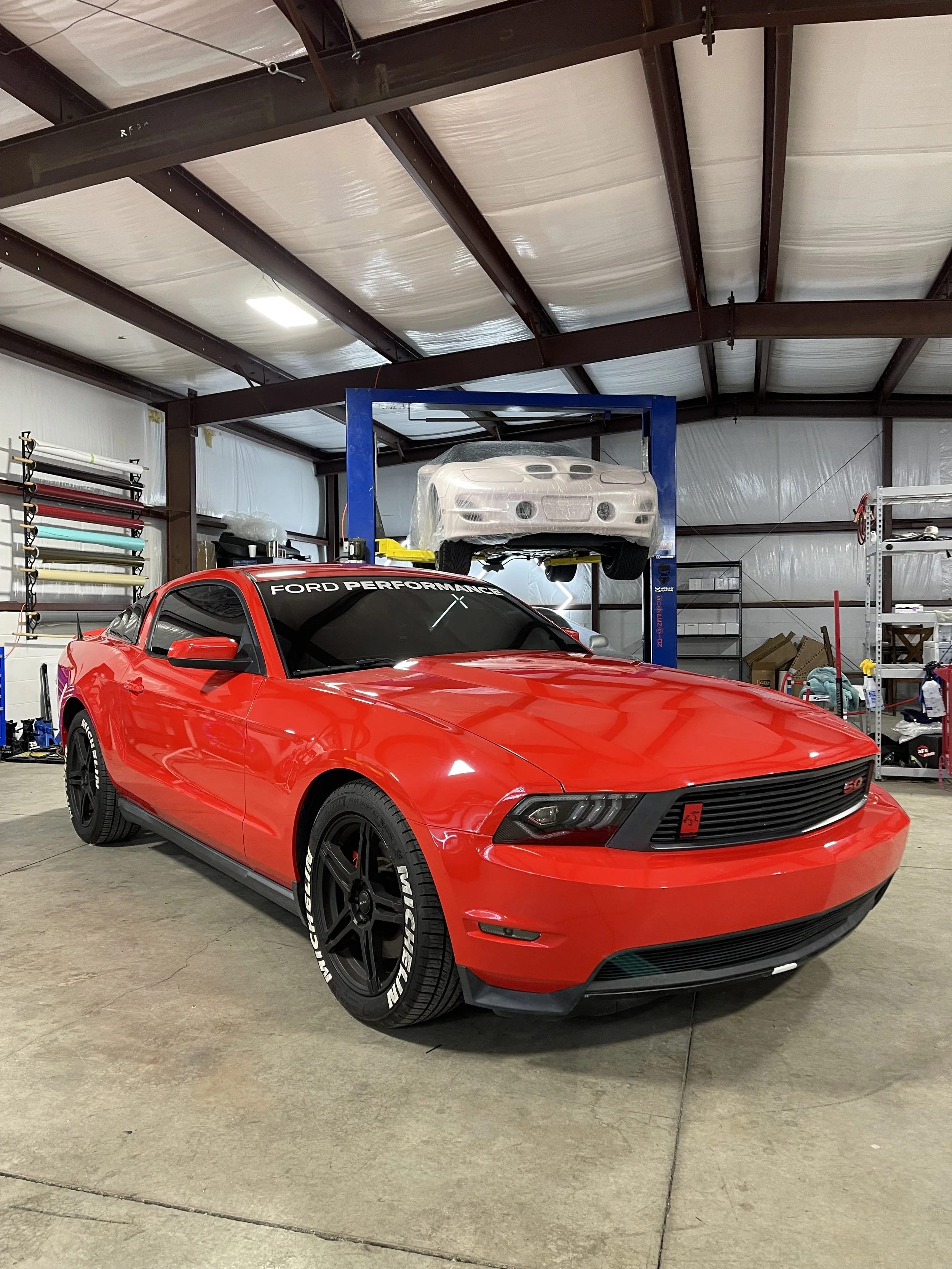Red Ford Mustang with black wheels and a "Ford Performance" windshield banner parked in a garage, a car lift with another car covered in plastic above it, and various tools and supplies on shelves.