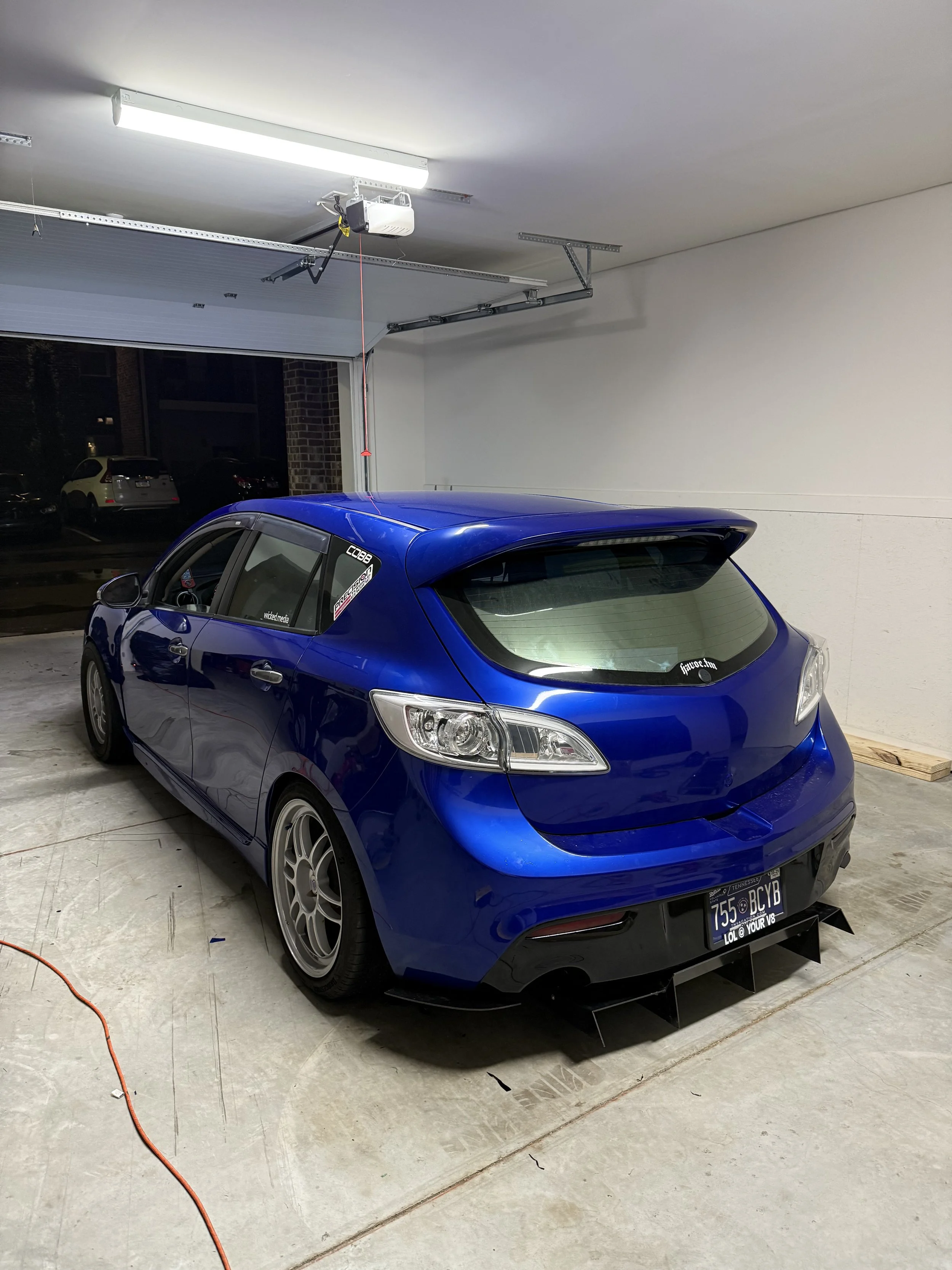 A blue hatchback car parked inside a garage with a raised and automatic garage door open. The car has aftermarket wheels and a rear spoiler, with stickers on the back window and side window.