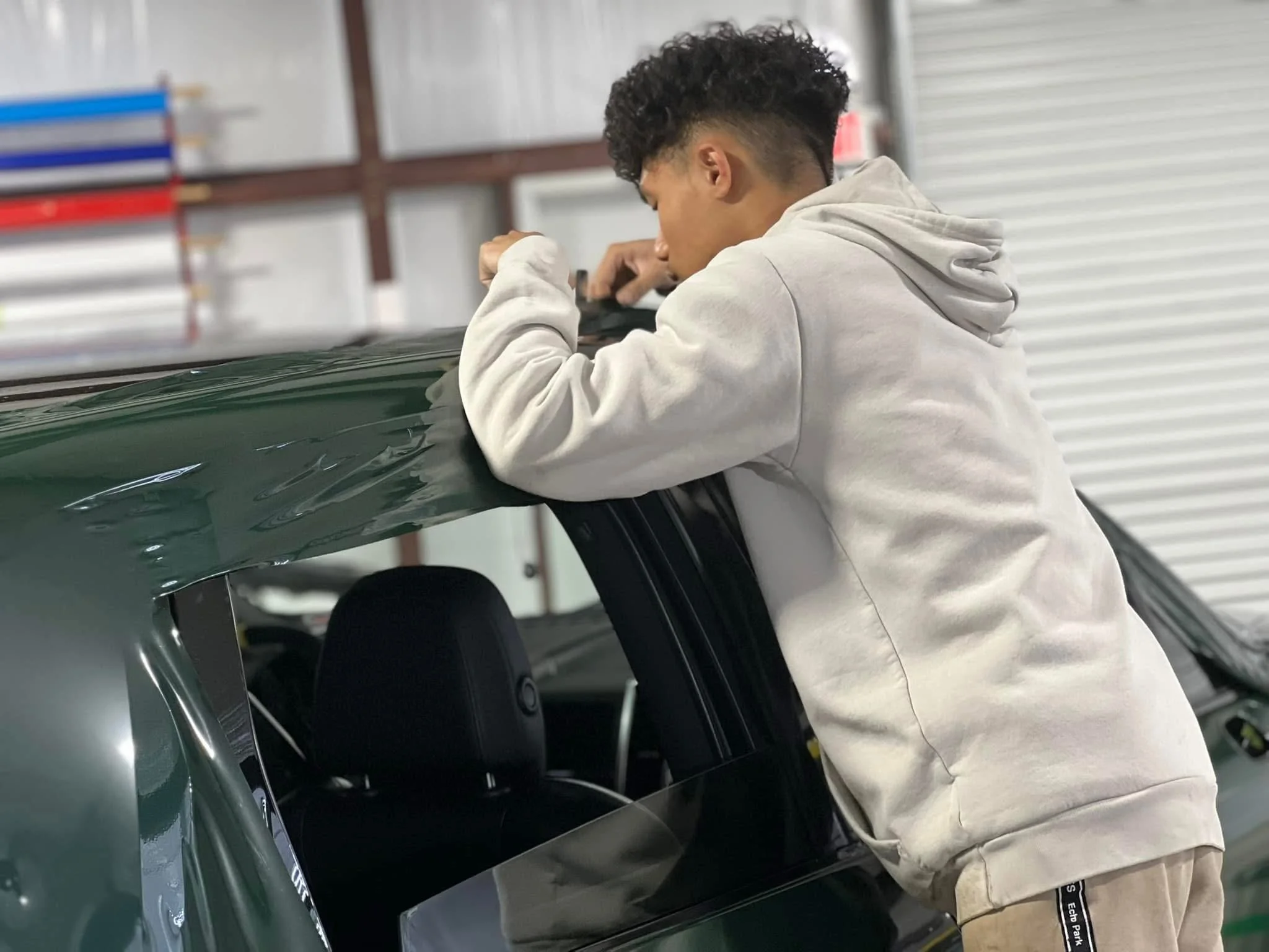 Young man working on a dark green car inside a garage, leaning over the car's roof with tools in hand.