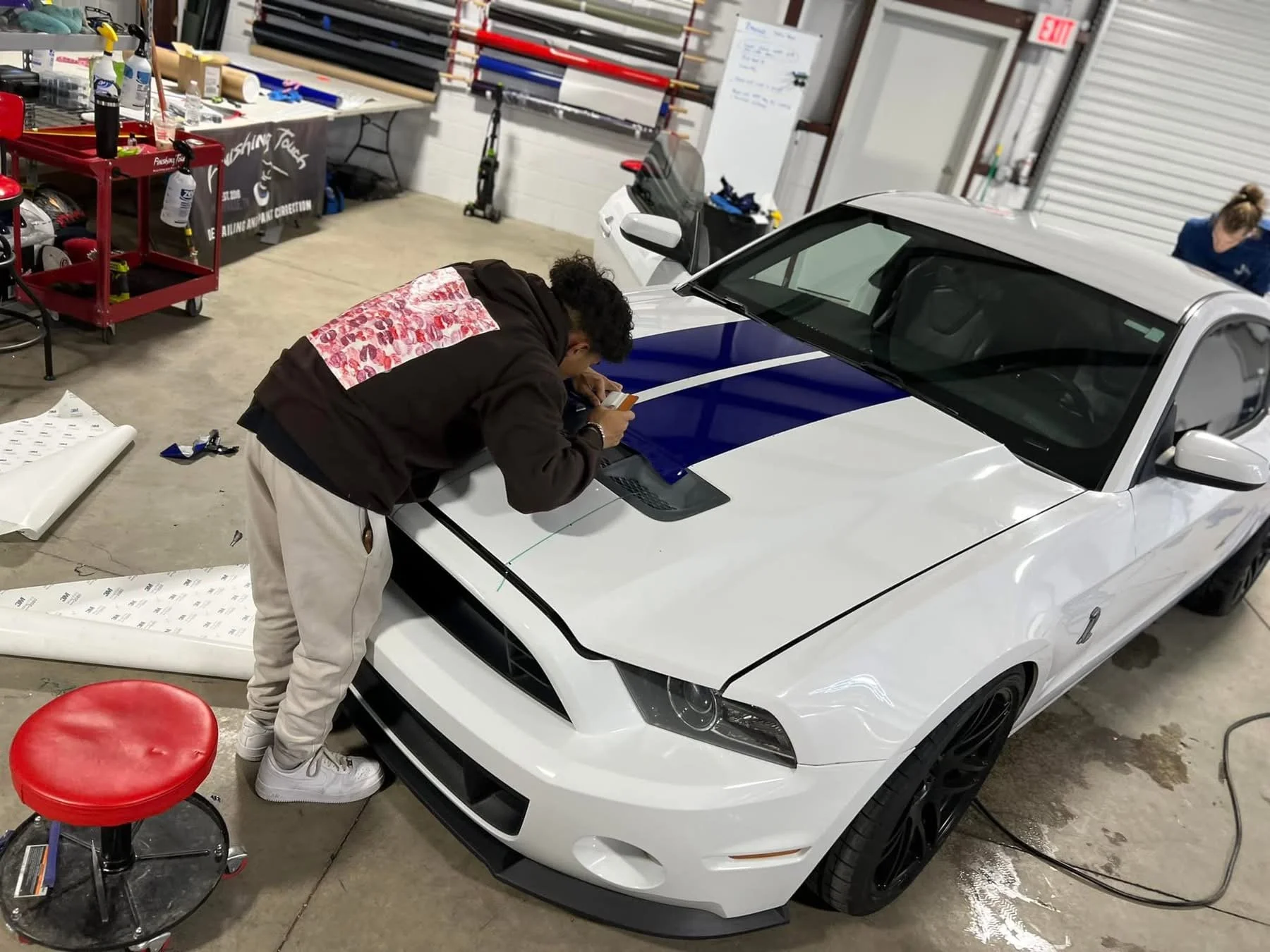A person in a black hoodie with red and white graphic on the back working on a white car inside a garage or workshop, with tools and equipment around. They are leaning over the car's hood, using a smartphone. The car has a blue stripe on the hood and