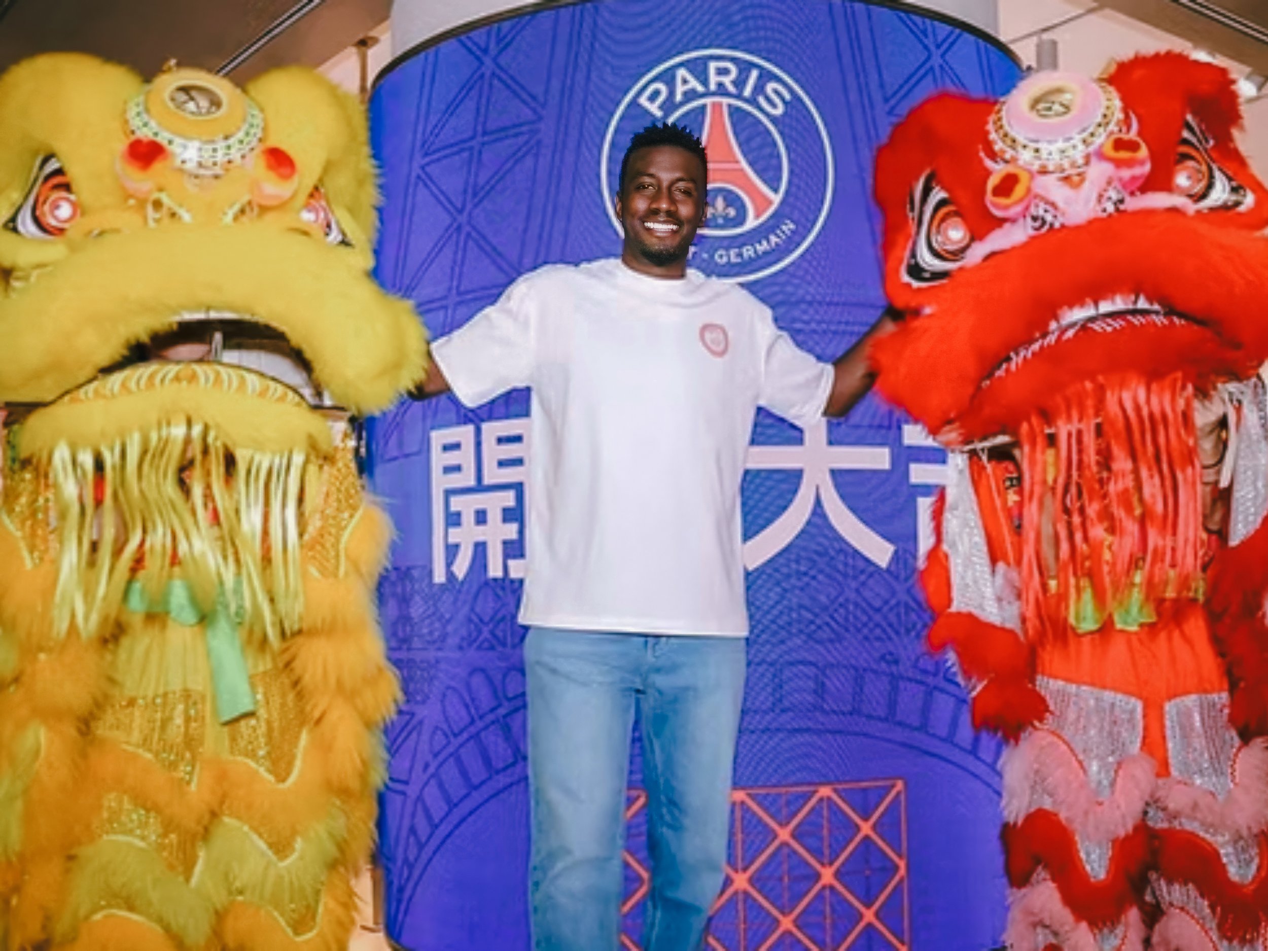 A person in a white T-shirt and blue jeans smiles while standing between two vibrant lion dance costumes, one yellow and one red, in front of a Paris Saint-Germain backdrop.