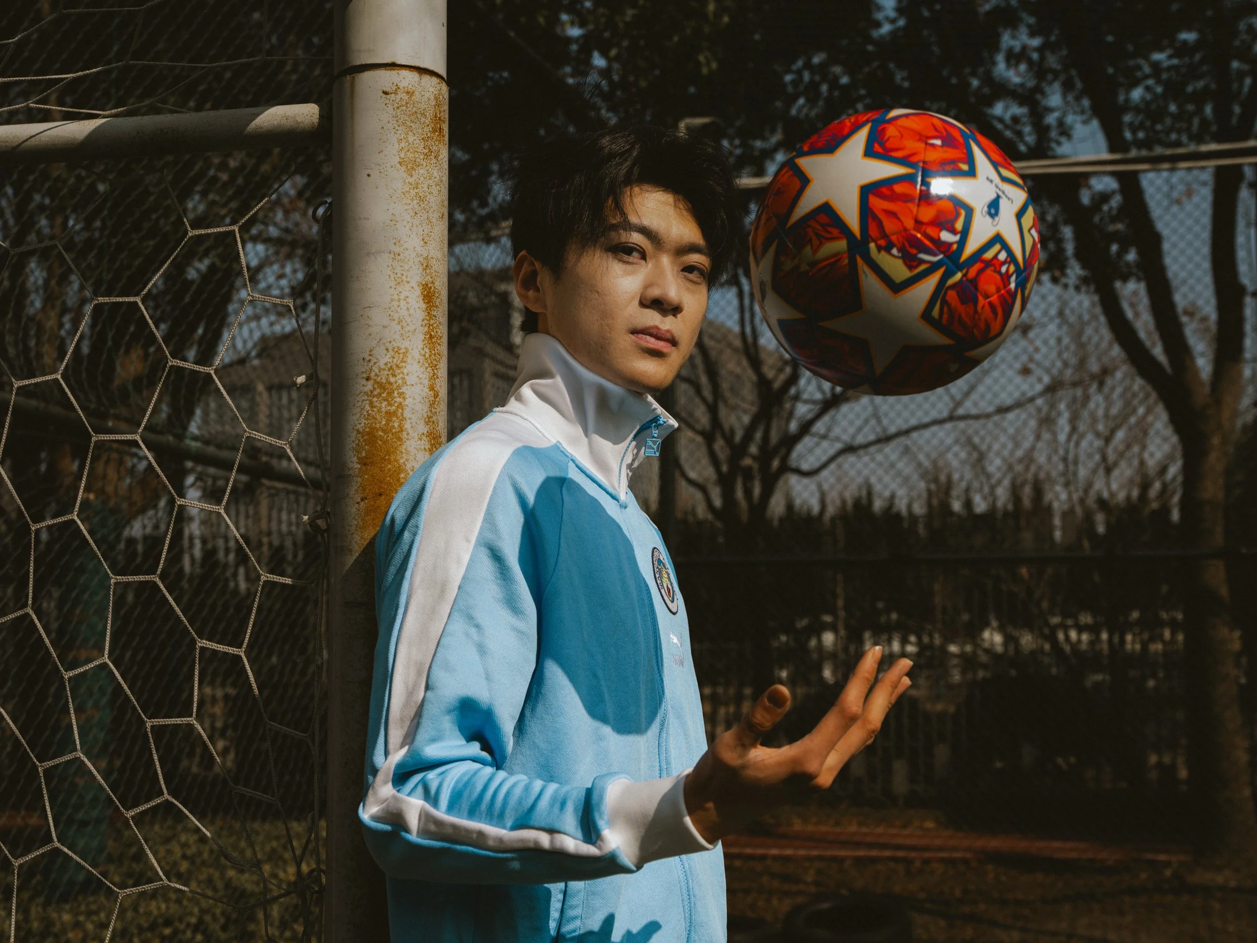 Young man in a blue jacket confidently tossing a colorful soccer ball near a goalpost.