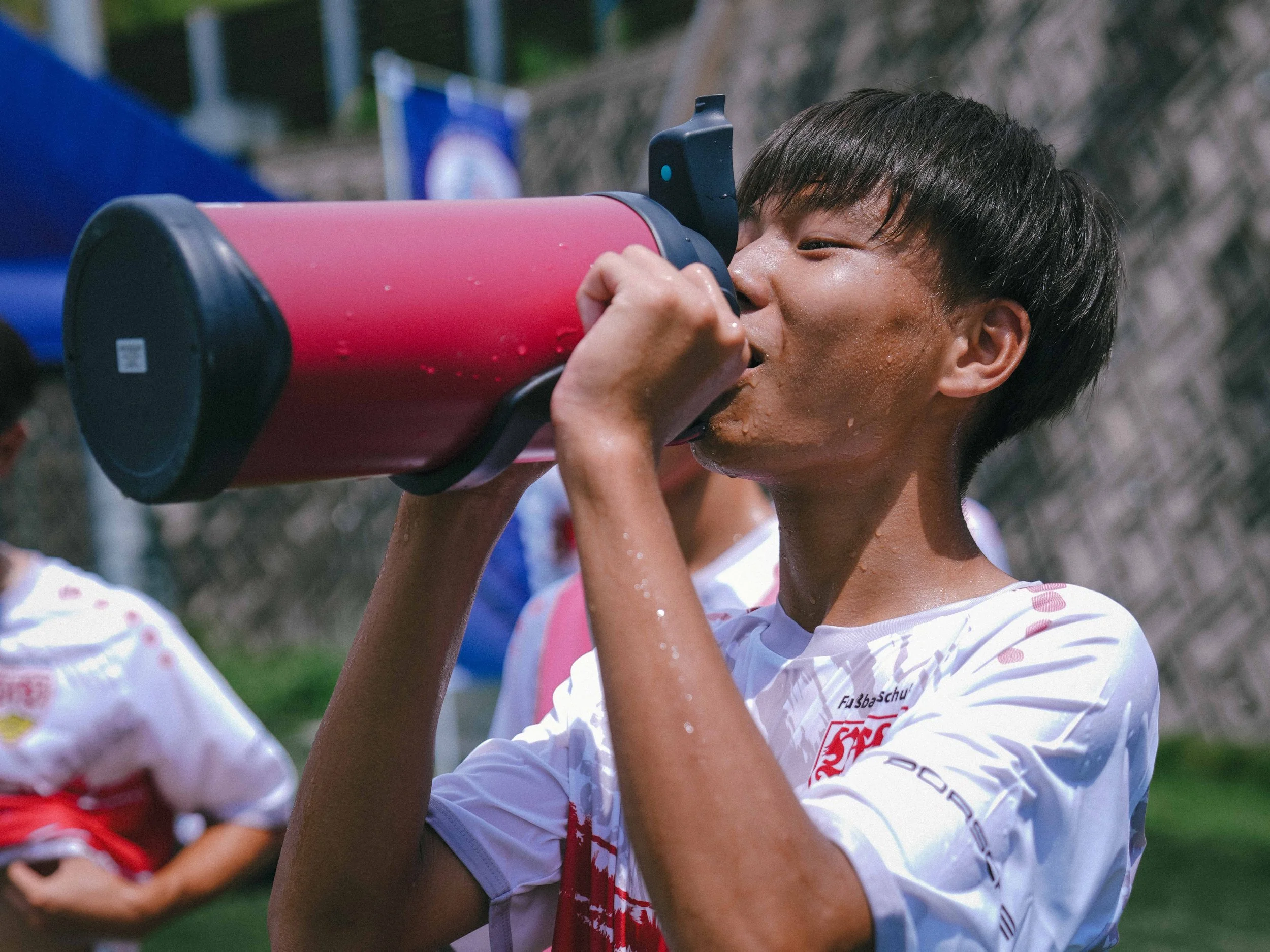 Young athlete in a white sports jersey drinks from a large red bottle, cooling down under the sunny sky. Energetic, refreshed, summer sports day.