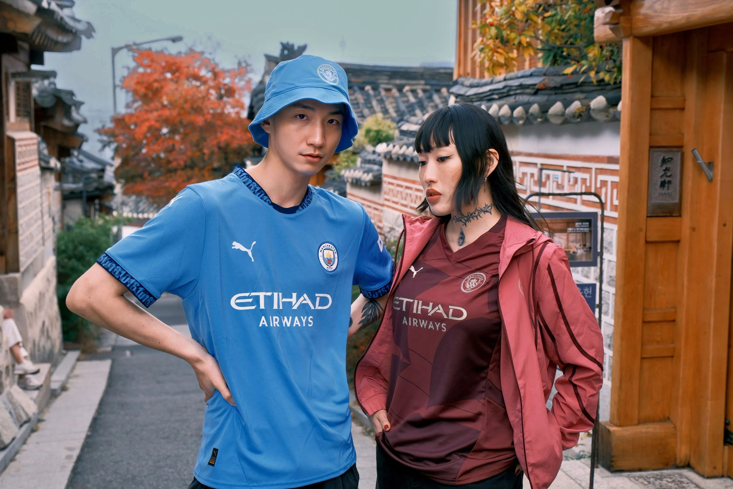 A young man and woman in sporty Manchester City and Manchester City jerseys standing outdoors in a traditional Asian setting with wooden structures and autumn foliage.