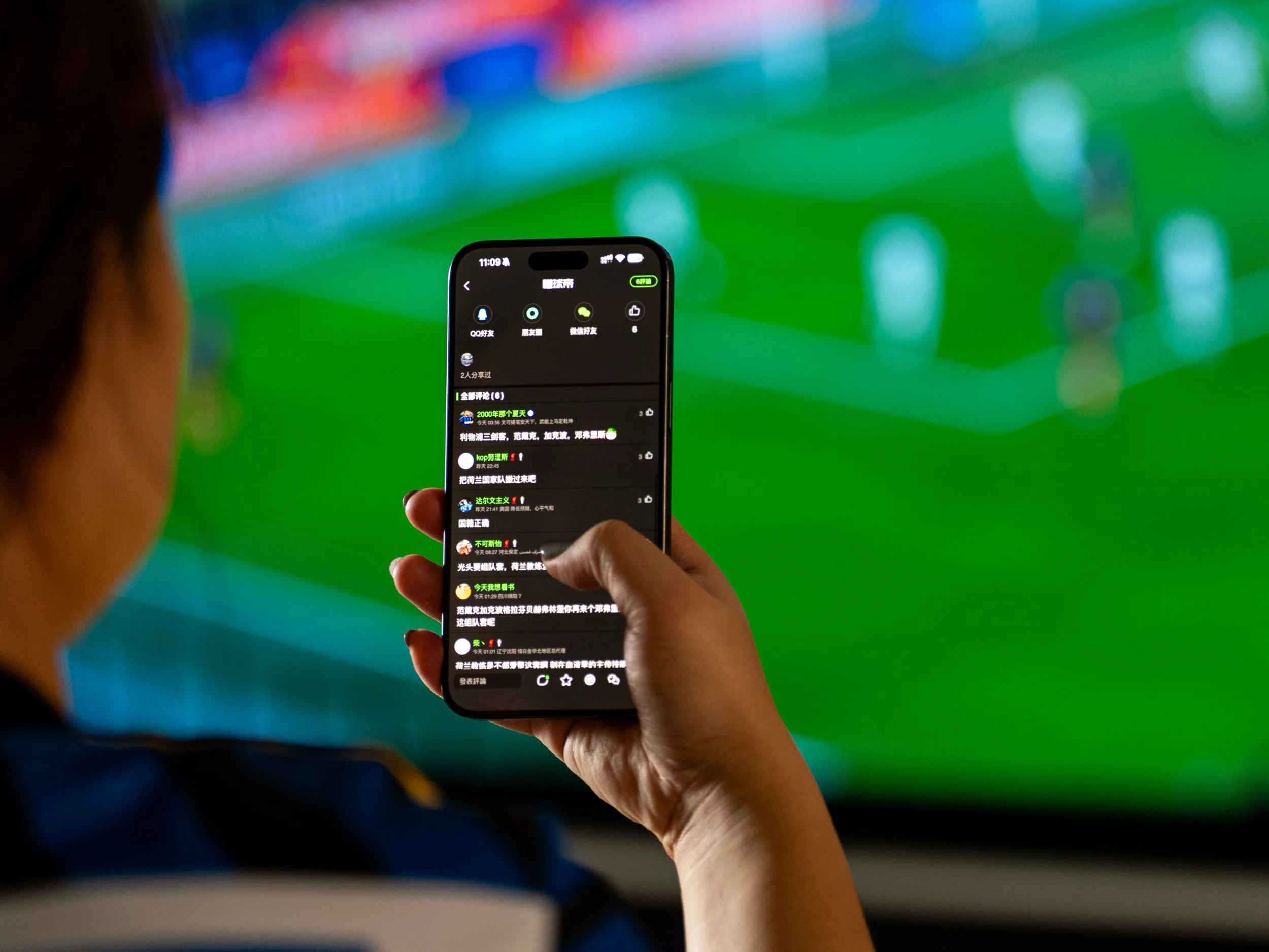 A person holds a smartphone with a messaging app open, showing text in Chinese. In the blurred background, a soccer match is on a TV screen, suggesting multitasking or real-time discussion.