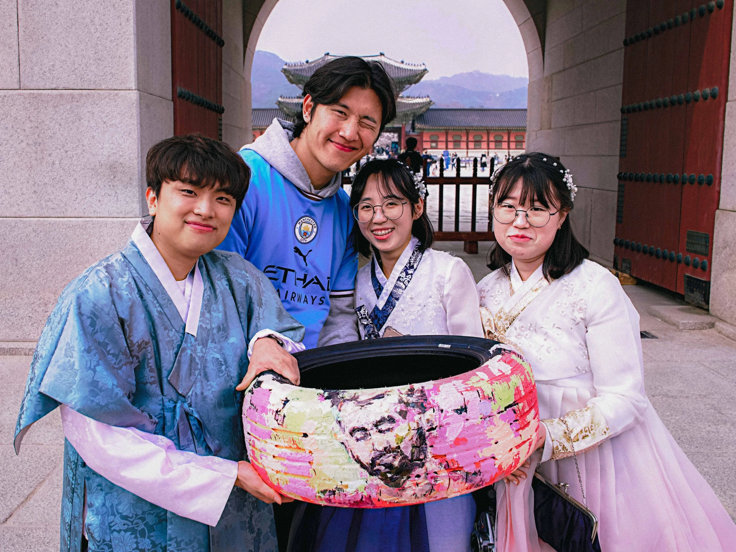 Four people stand smiling in traditional Korean hanboks and a sports jersey, holding a colorful painted tire.