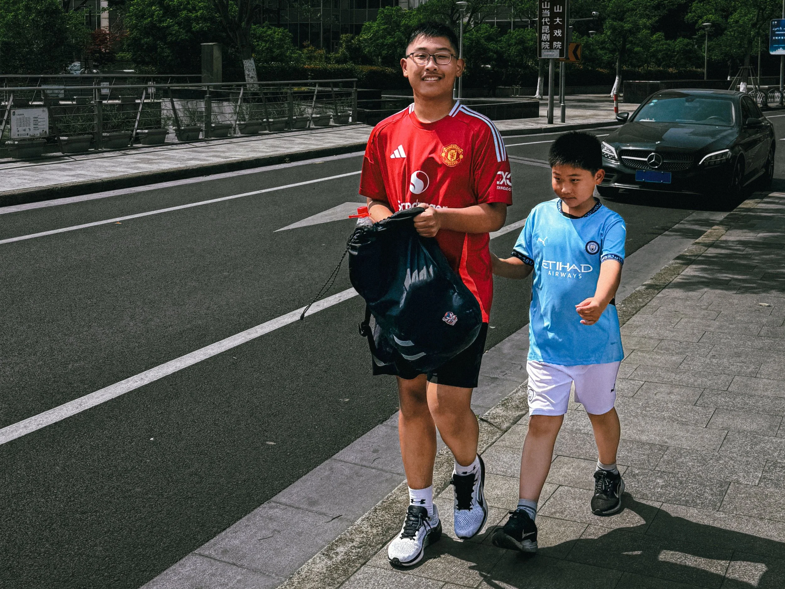 Two Chinese boys walking along a city street, one in a red sports jersey and the other in a blue one.