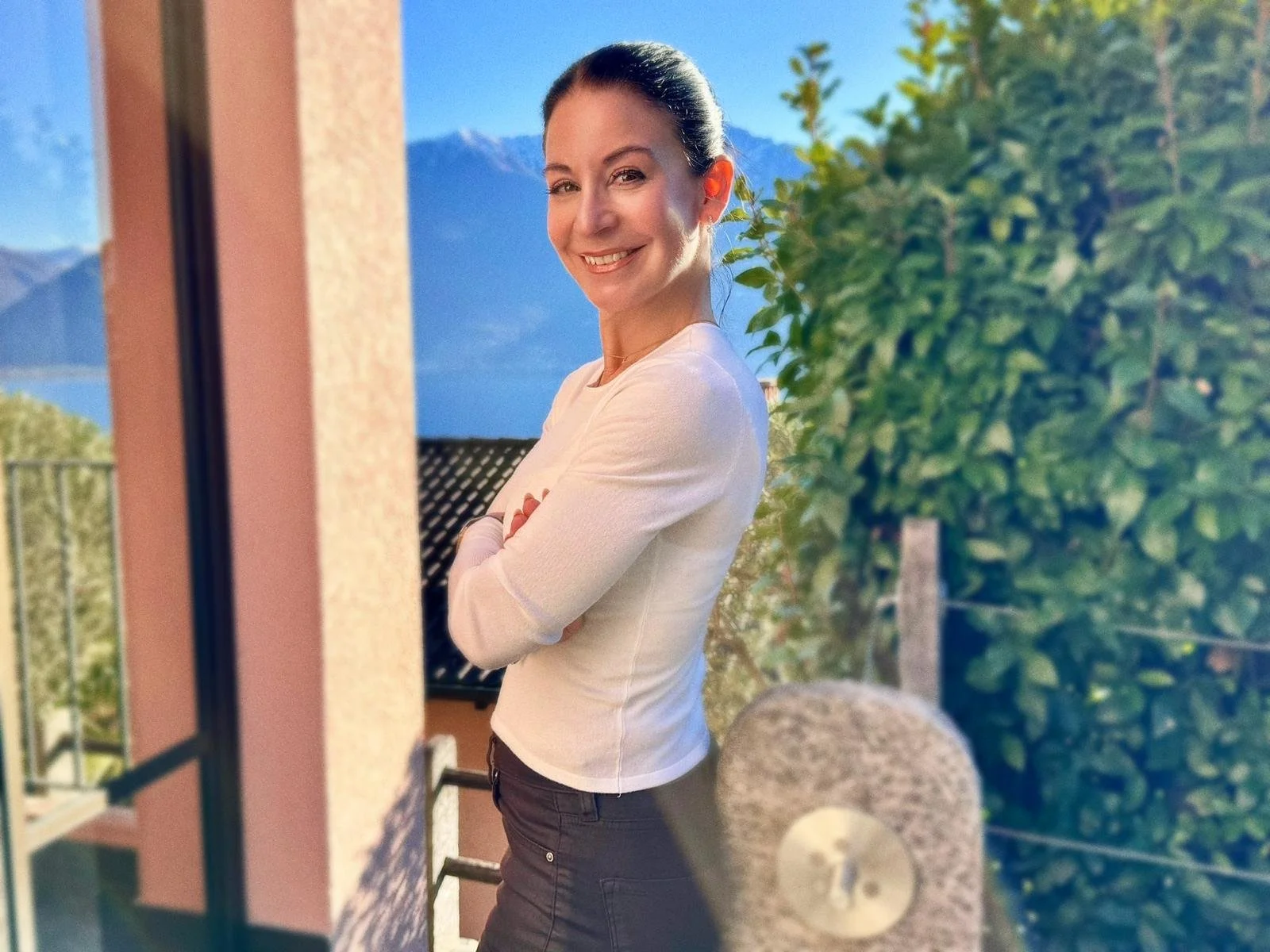 A woman with dark hair tied back, wearing a white long-sleeve top and black pants, standing on a balcony with a mountain landscape in the background, smiling at the camera.