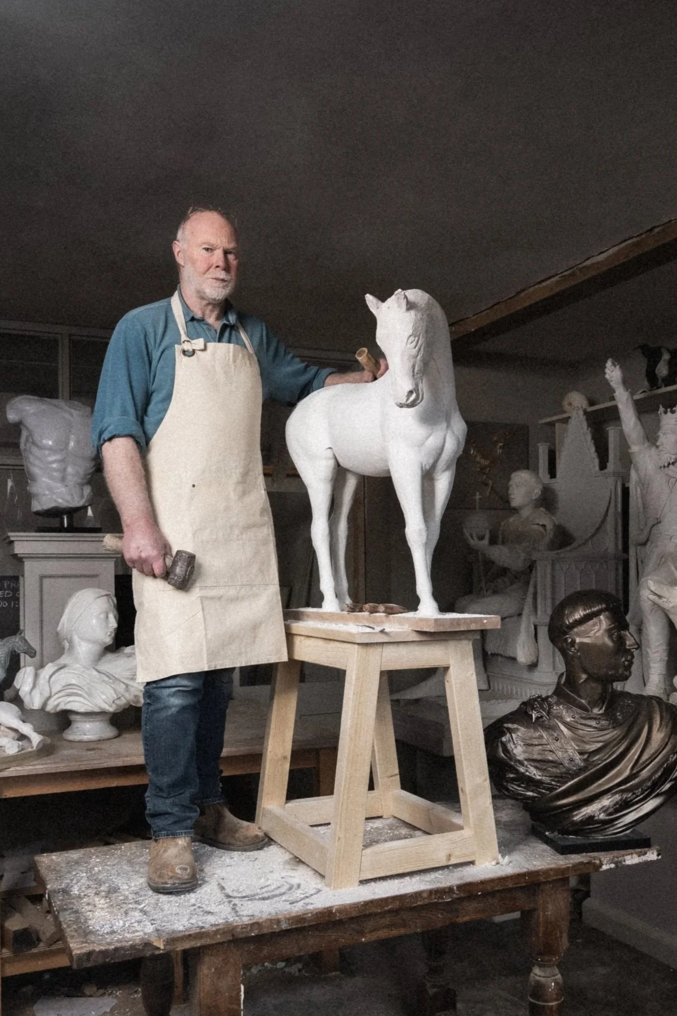 An older man with a beard wearing a blue shirt, apron, and boots is sculpting a white horse sculpture in an art studio. The man is holding a sculpting tool, and the horse sculpture is on a wooden stand. The studio has various busts and sculptures in the background.