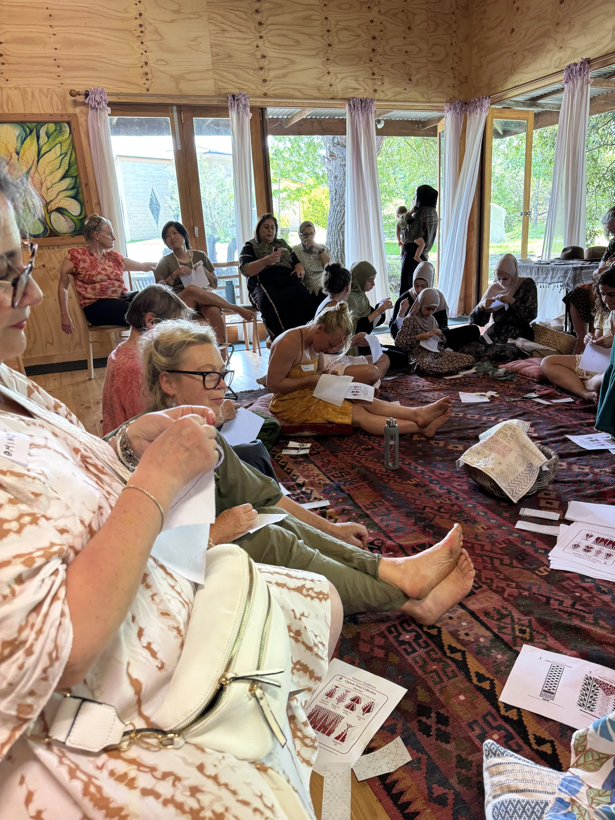Group of women and girls seated on a colorful rug inside a wooden room, engaging in a workshop or class, with printed materials and papers around.