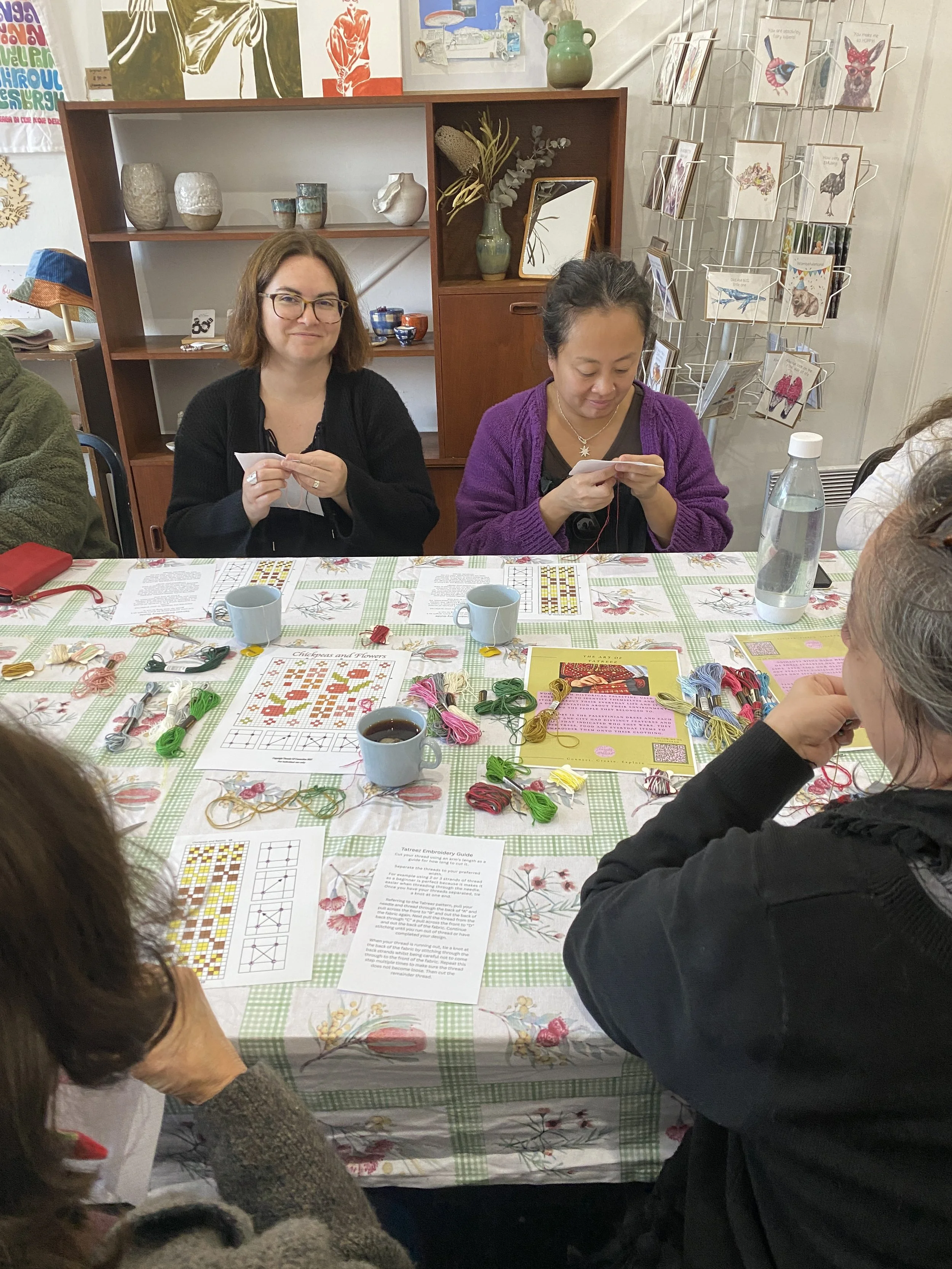 A group of women sitting around a table with embroidery supplies, including colorful threads, patterns, and cups of coffee, engaging in a craft workshop.