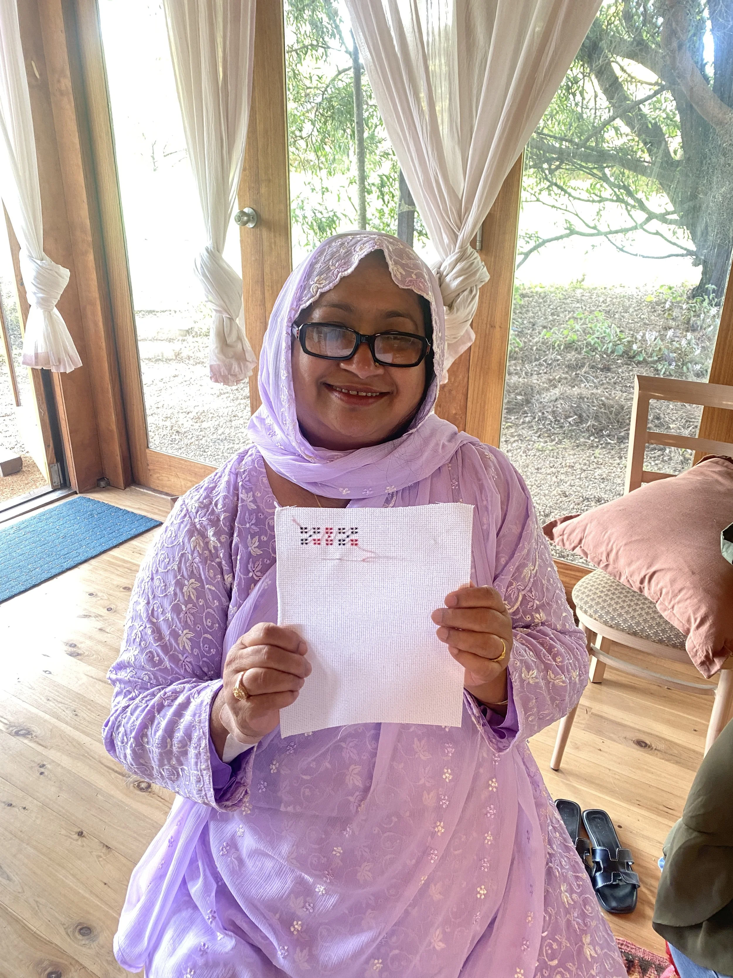 A woman wearing glasses and a pink lace dress with a matching headscarf, holding a piece of white cross-stitched fabric with red and black thread, smiling indoors near glass doors with white curtains, and a view of trees outside.
