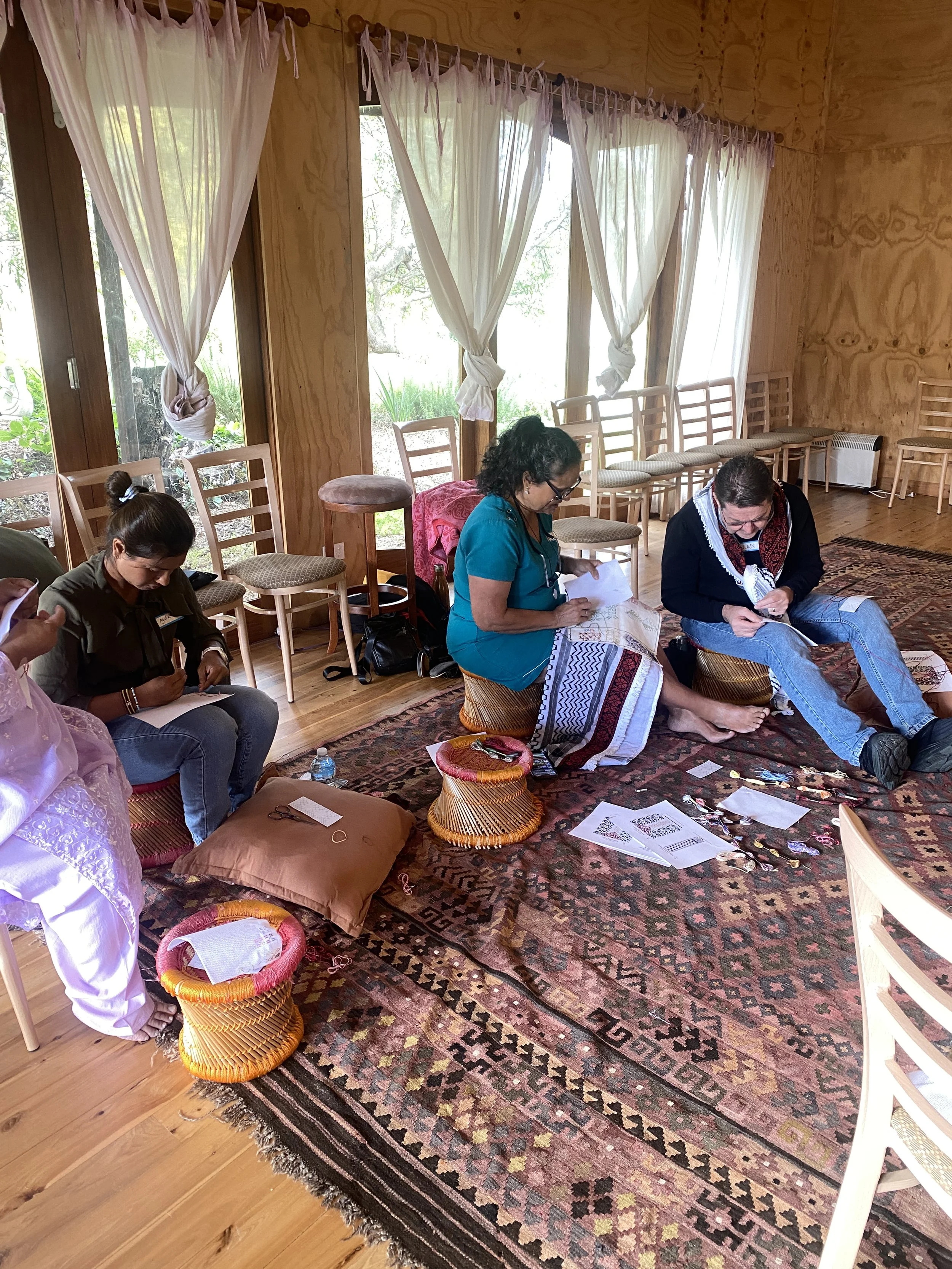 Group of women sitting on cushions on a patterned rug, engaging in handcraft activities indoors near large windows with white curtains.