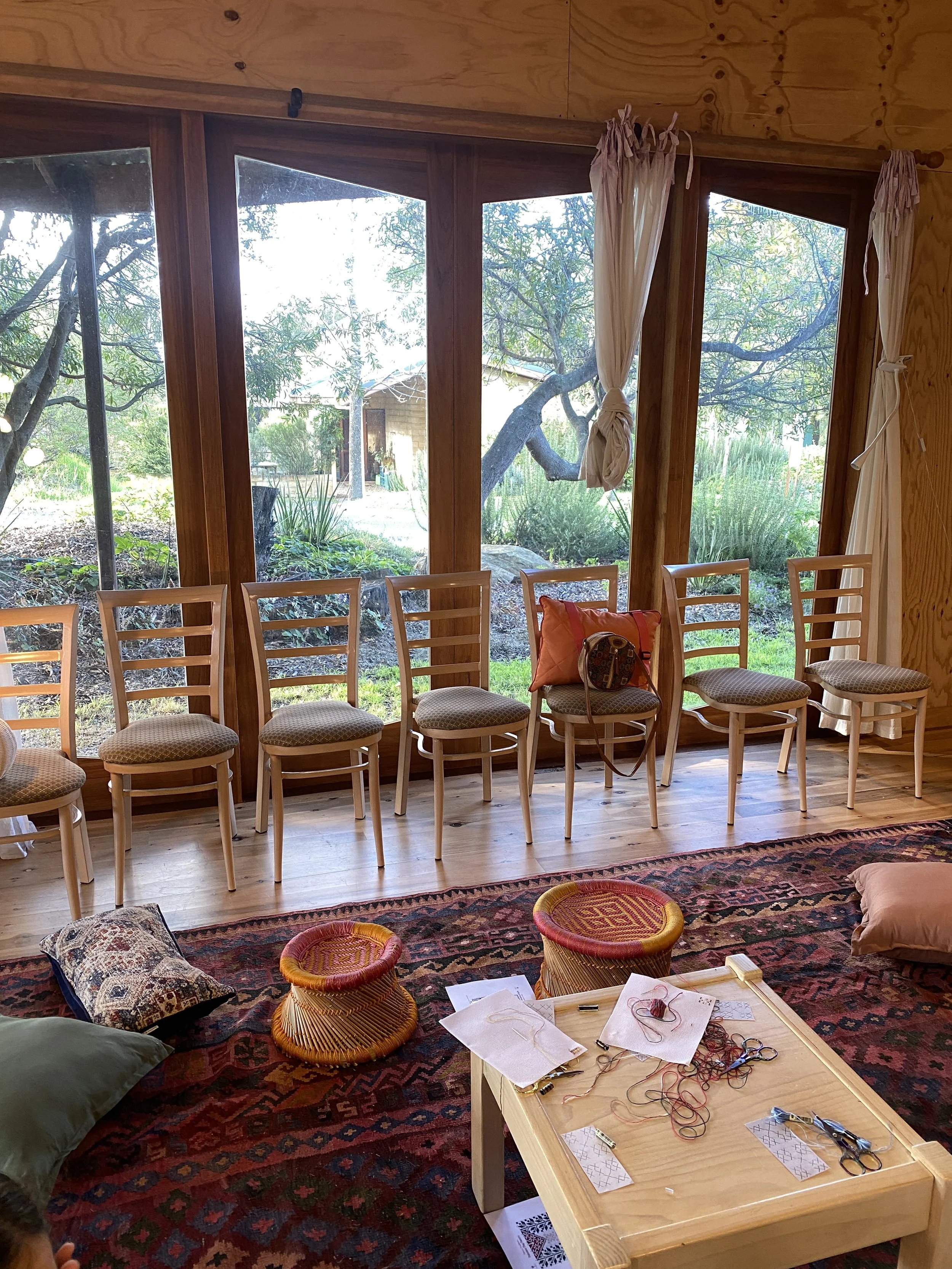 A room with a large window showing trees and outdoor scenery. Several wooden chairs are arranged in a row against the window. On the floor, there are two small woven stools, pillows, and a wooden table with embroidery supplies and scissors.