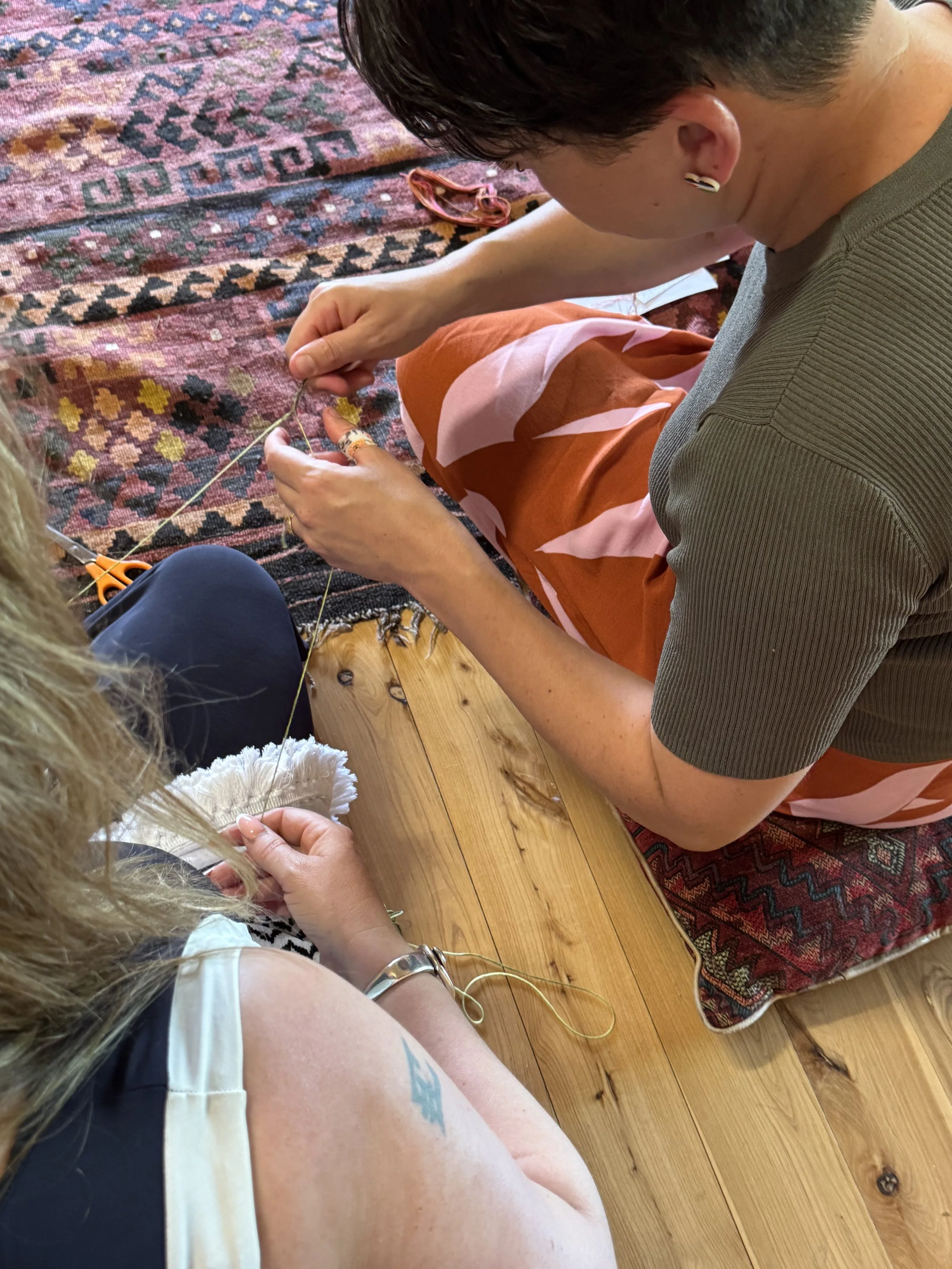 Two women sit on the floor, engaging in a craft activity involving thread and fabric. The woman on the right holds a needle, and the woman on the left holds a piece of fabric. There are scissors on the floor, and they are sitting on a wooden floor wi