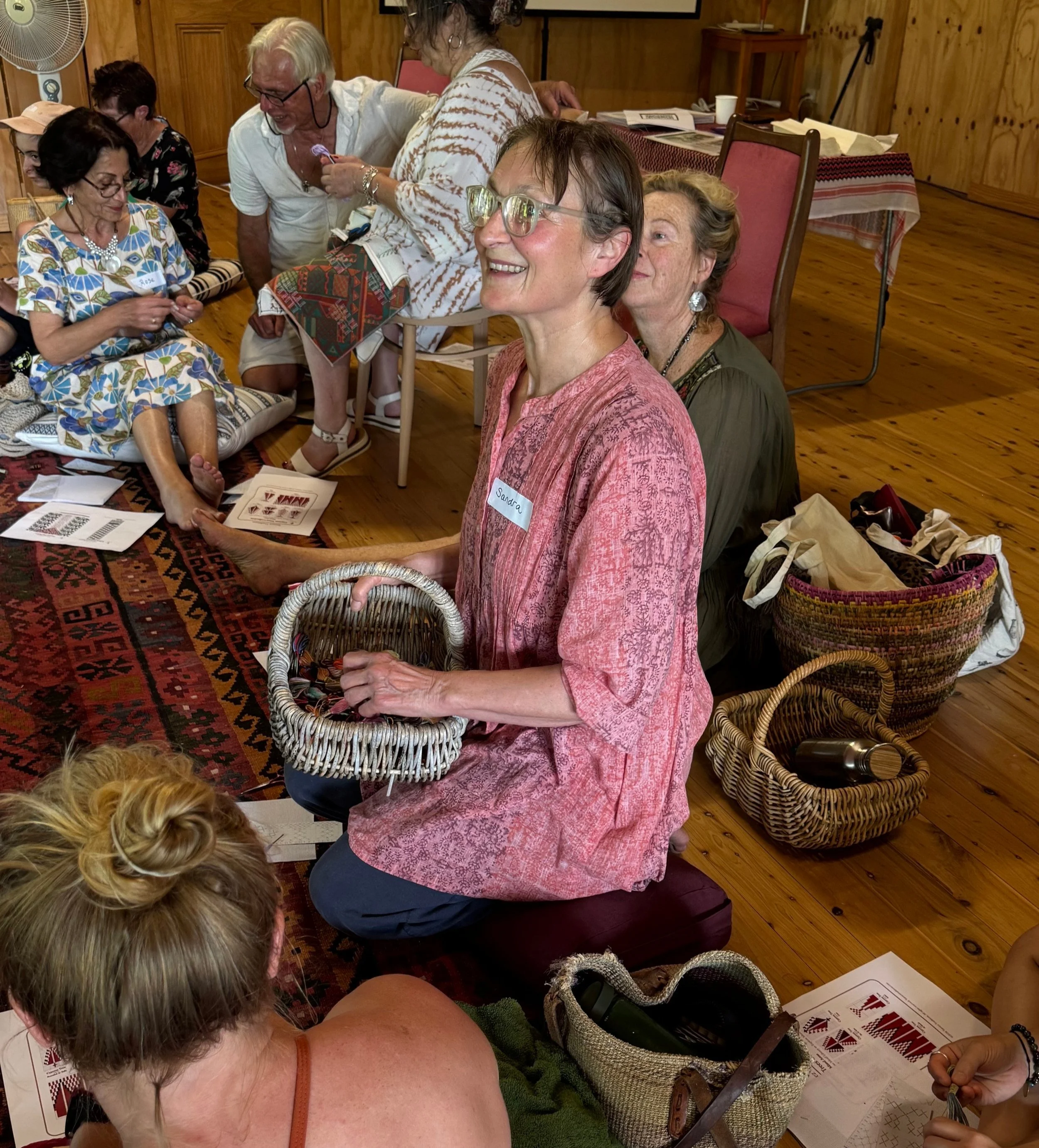 A group of people sitting on a colorful rug in a cozy wooden room participating in a craft or workshop activity. The woman in the foreground is smiling, holding a small woven basket, and wearing glasses and a pink patterned top. The scene includes baskets, papers, and supplies around them.