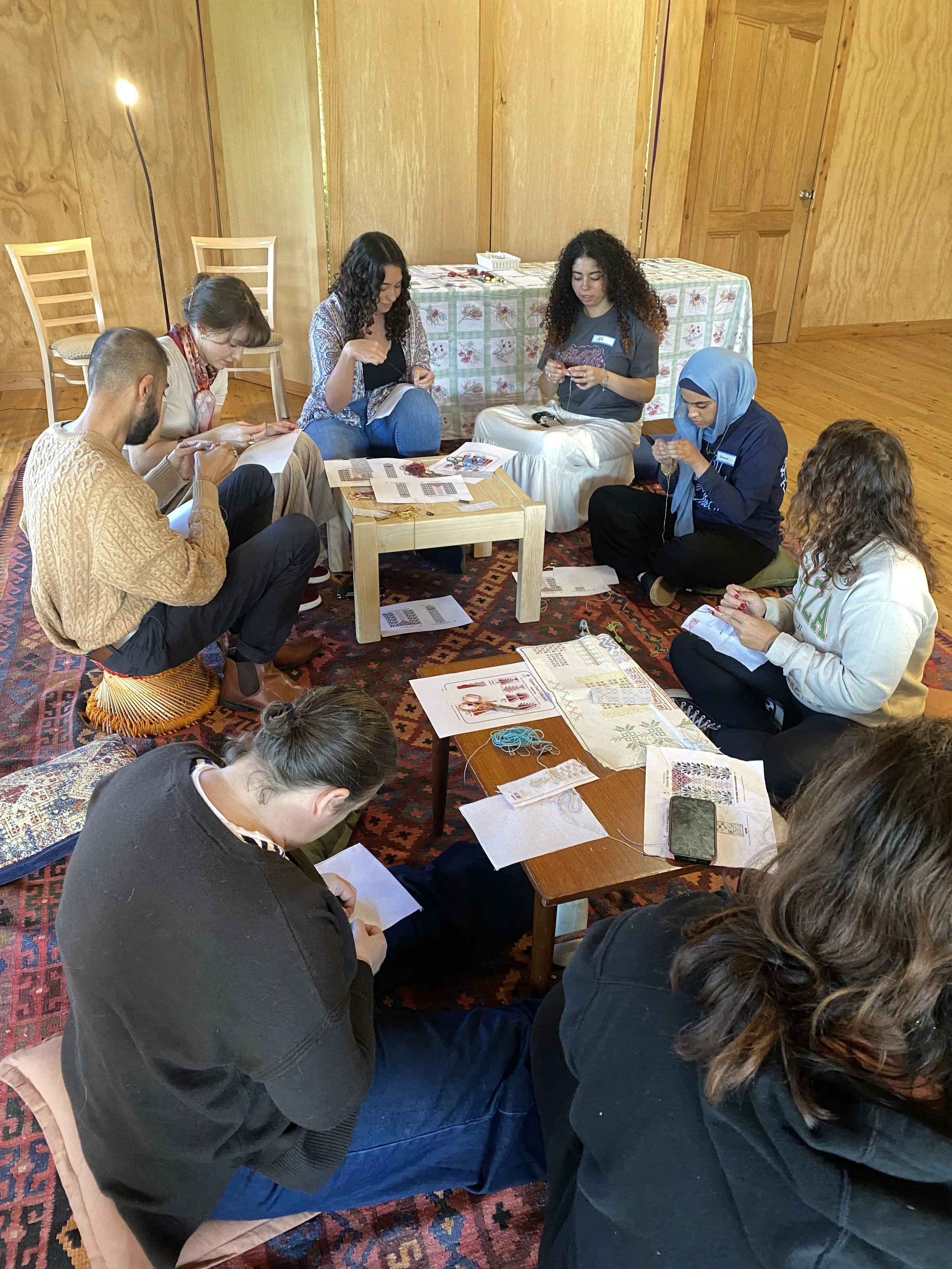 Group of people sitting on the floor and chairs, engaged in a crafting or knitting activity in a cozy wooden room.