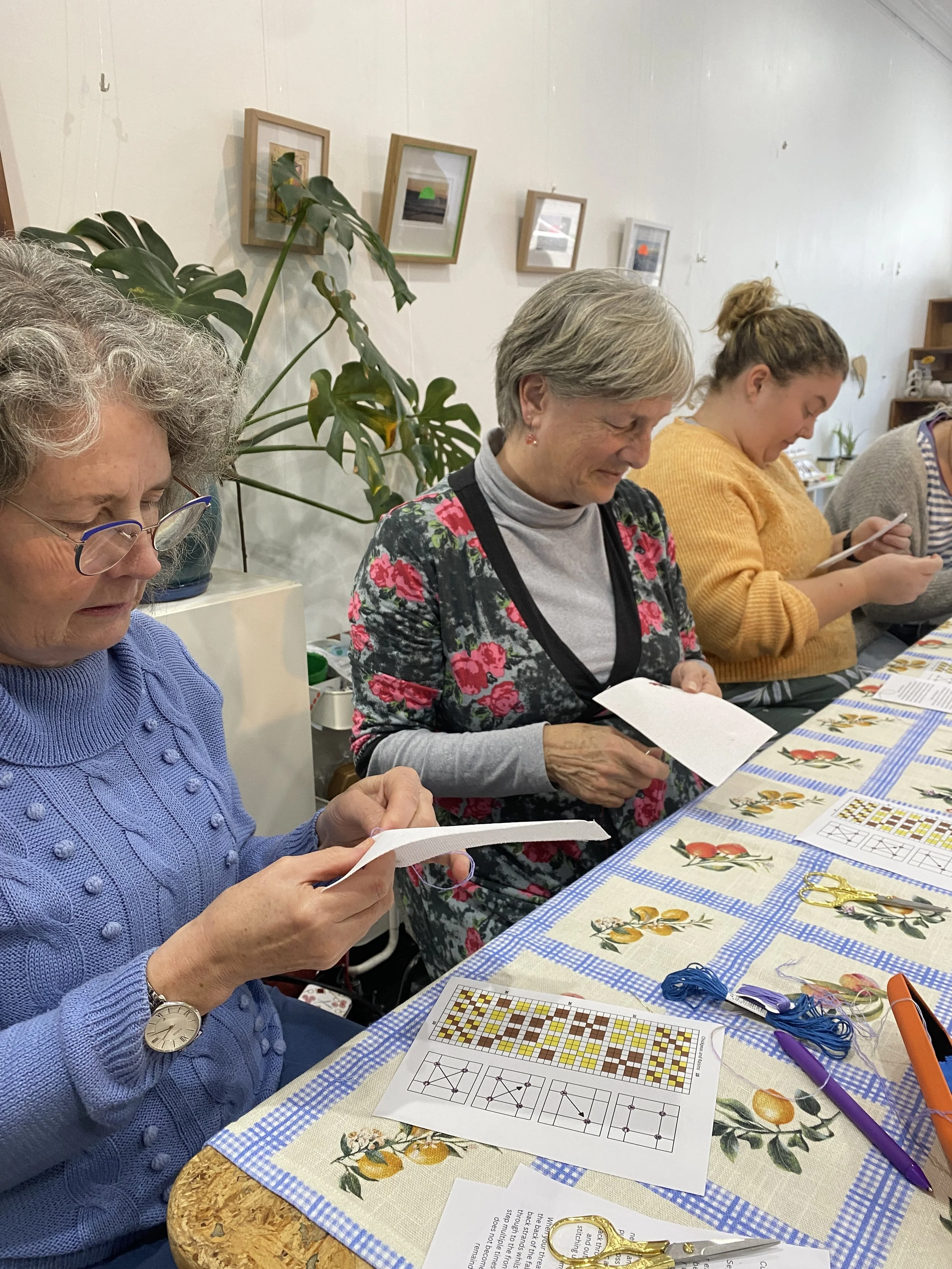 Four women sitting at a table with craft supplies, reading papers, engaged in a craft activity; the table is covered with a lemon-themed tablecloth and there are small framed pictures on the wall behind them.
