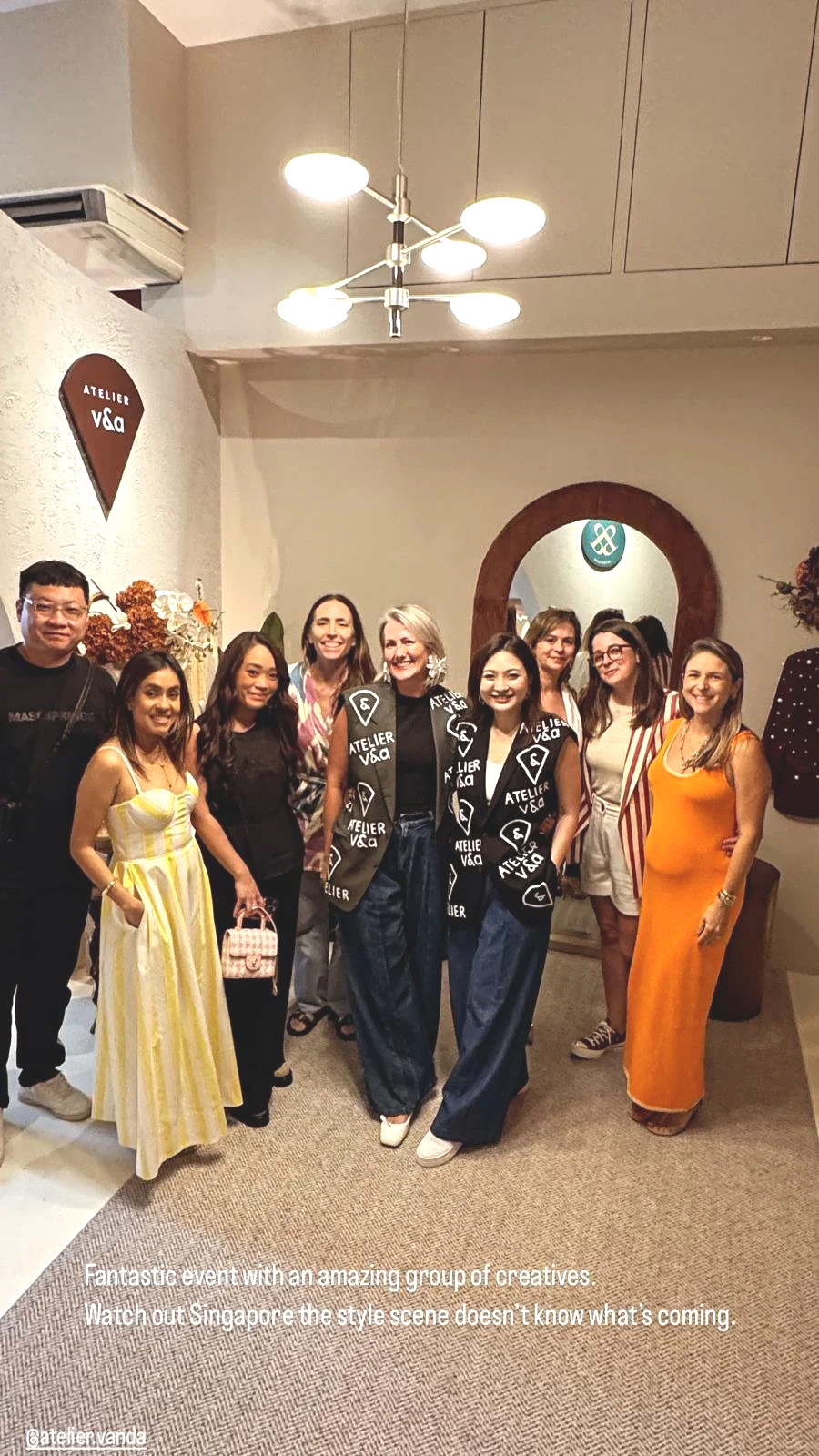 Group of eight women and one man standing together in an indoor setting, smiling for the photo. They are dressed in colorful and stylish clothing. The background features a mirror, floral arrangements, and a wall decor sign that says 'Atelier v&d'.