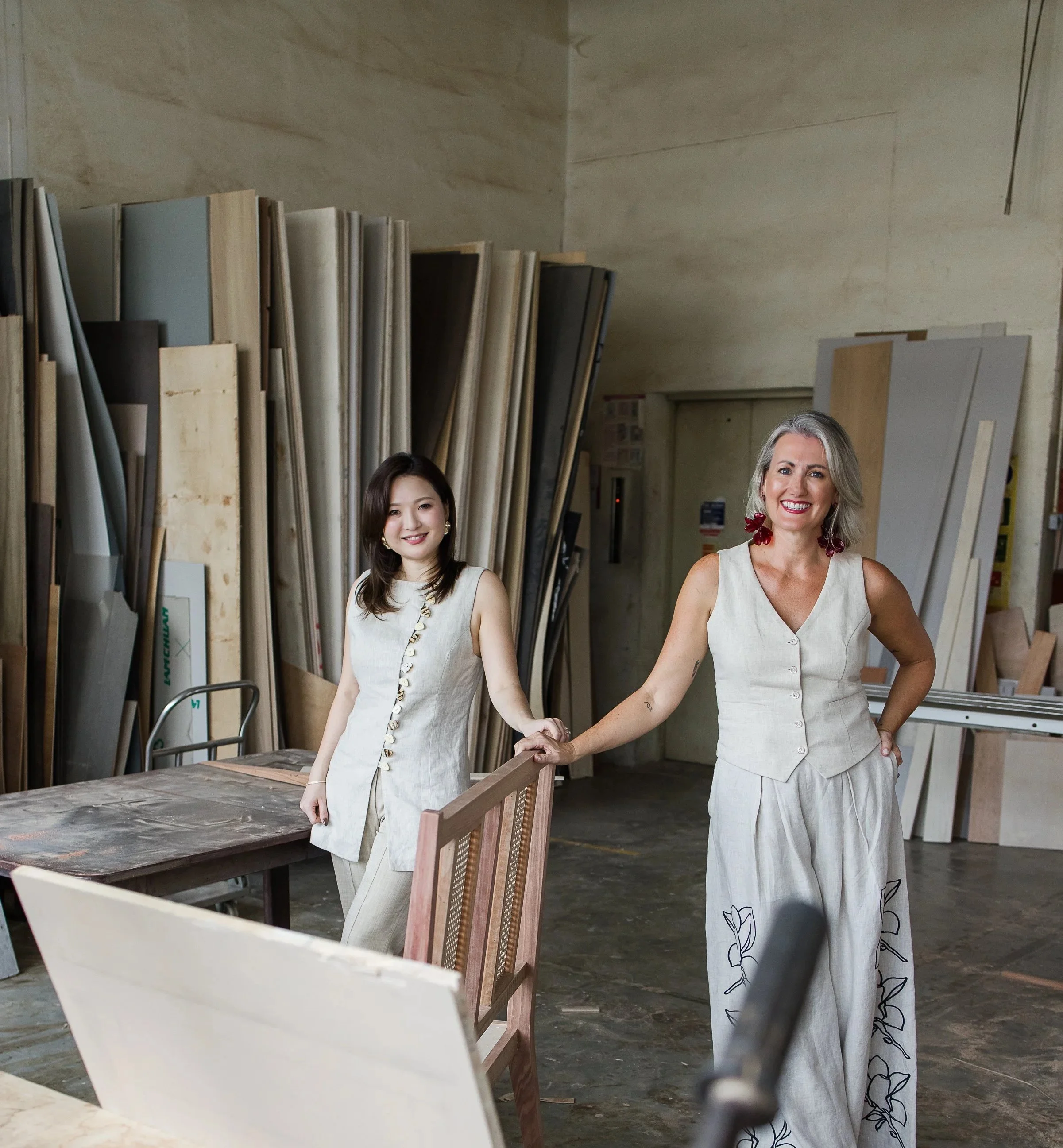 Two women holding hands in a woodworking or furniture workshop, surrounded by woodworking materials and tools, smiling at the camera.