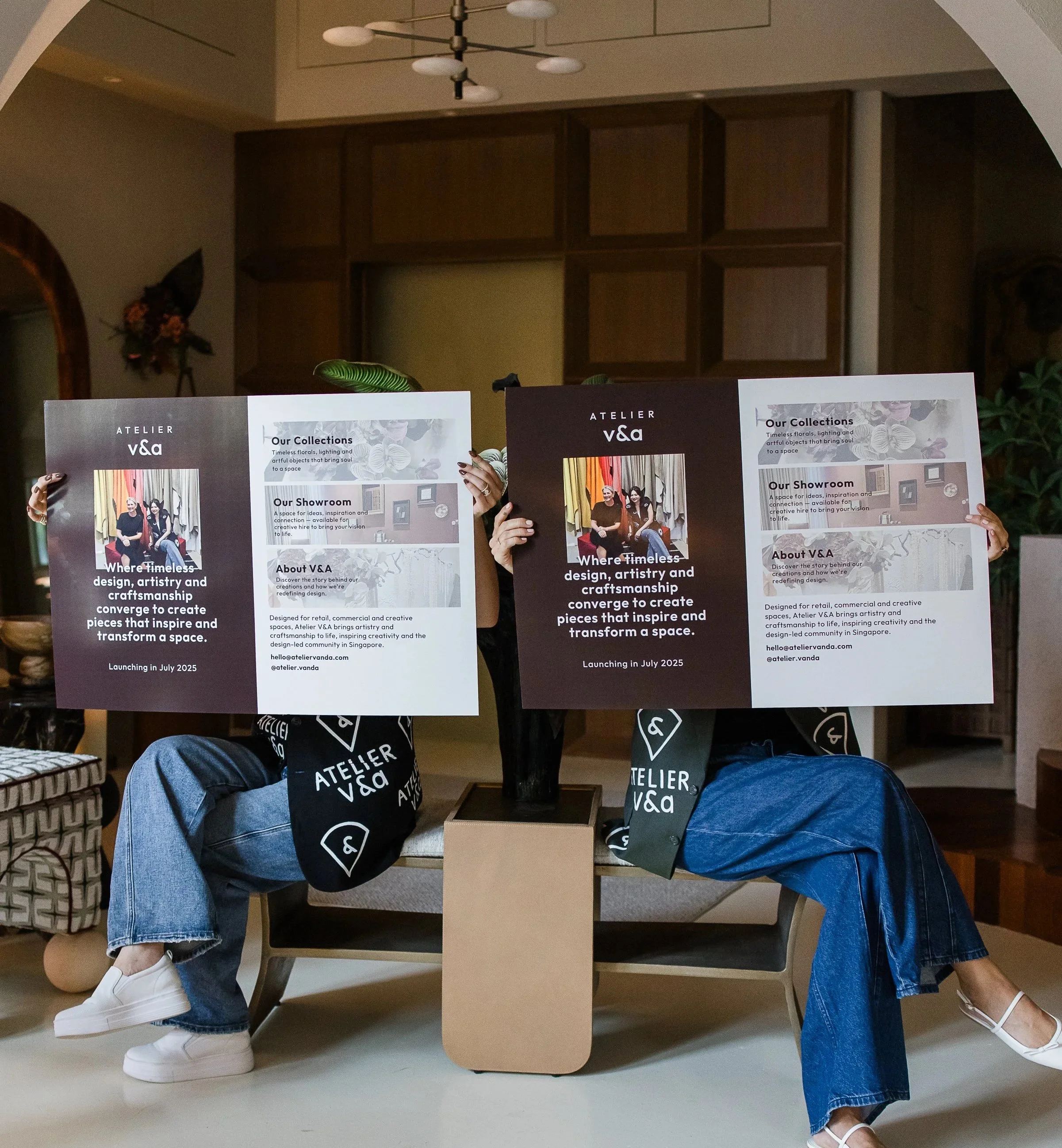 Two people sitting on a bench behind a table, holding large signs promoting Atelier V&A, a design and craftsmanship community. The signs contain text about collections, showroom, and the community's mission to inspire and transform spaces. The people are wearing black and blue jeans, with their faces obscured by the signs.