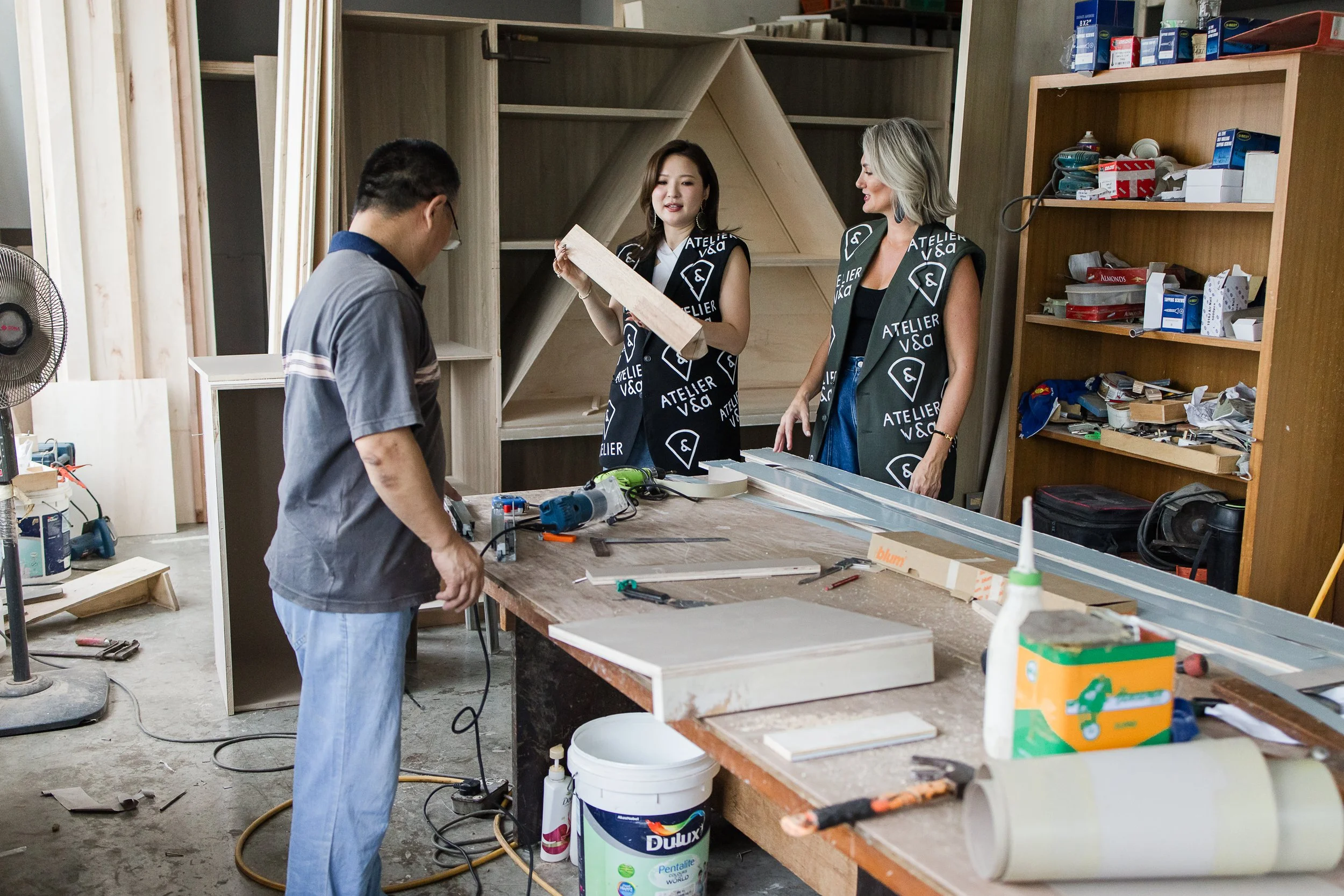Two women and one man working in a woodworking shop. The women are holding a piece of wood, and the man is looking down at the workbench, which is cluttered with tools and materials. The women are wearing matching black vests with the logo 'Atelier V&A'.
