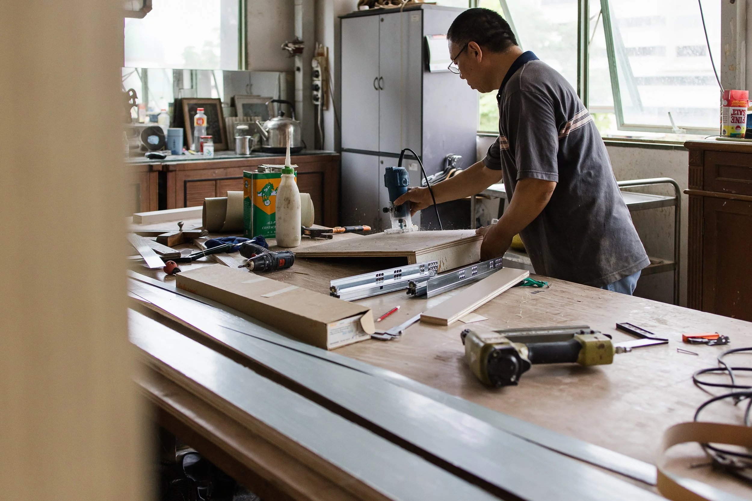 A man working in a workshop, using a jigsaw to cut a piece of wood. The workbench is cluttered with tools, wood pieces, and supplies, with a window in the background providing natural light.