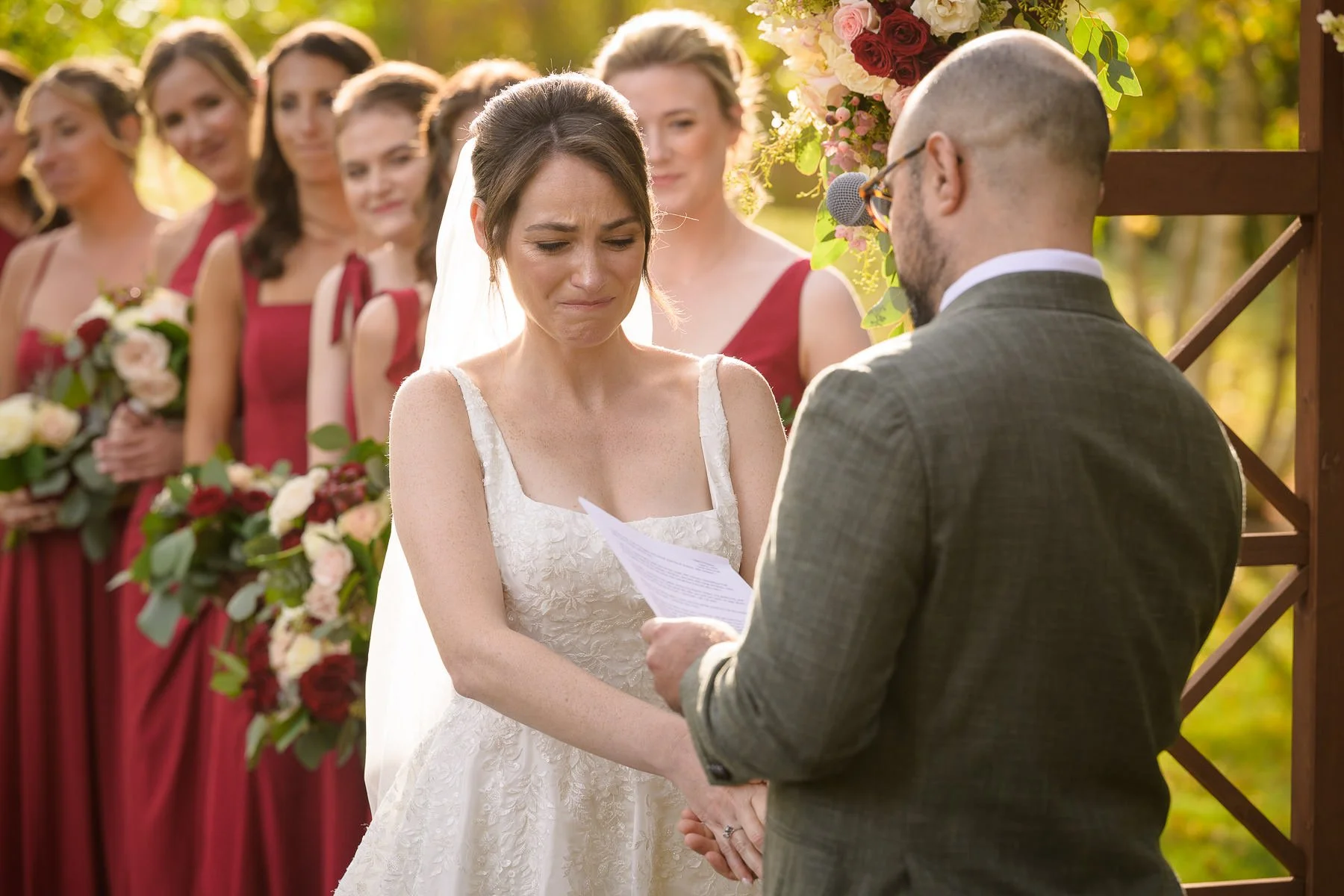 Chelsea becomes emotional while listening to David’s vows during their outdoor wedding ceremony at the Inn at Manchester in Vermont. Bridesmaids in red dresses stand behind her as the couple holds hands beneath a floral arbor.