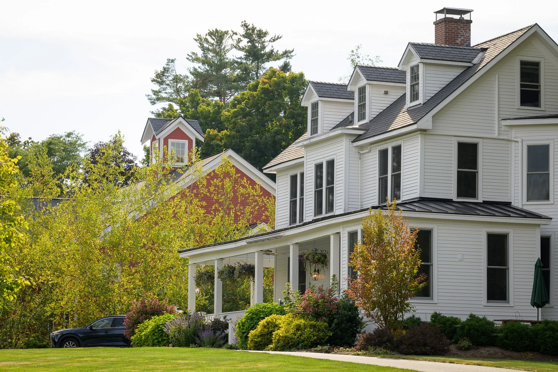 Exterior view of the Inn at Manchester in Vermont, with the white inn in the foreground and the red barn visible behind it. Trees, landscaping, and the open lawn frame this Manchester wedding venue in soft daylight.