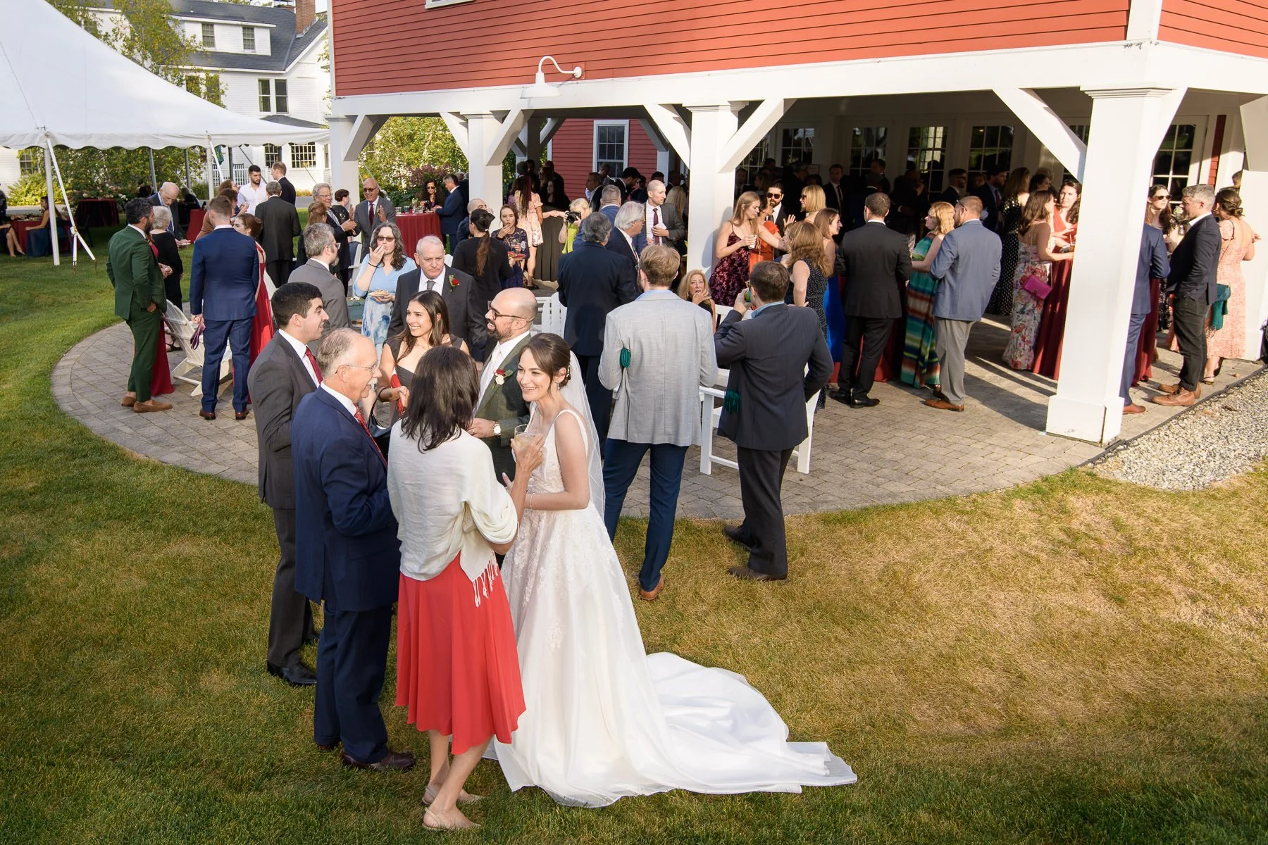 Guests gather for cocktail hour outside the red barn at the Inn at Manchester in Vermont as Chelsea and David mingle with family and friends on the lawn and patio. The wide scene shows the welcoming, social atmosphere of the reception.
