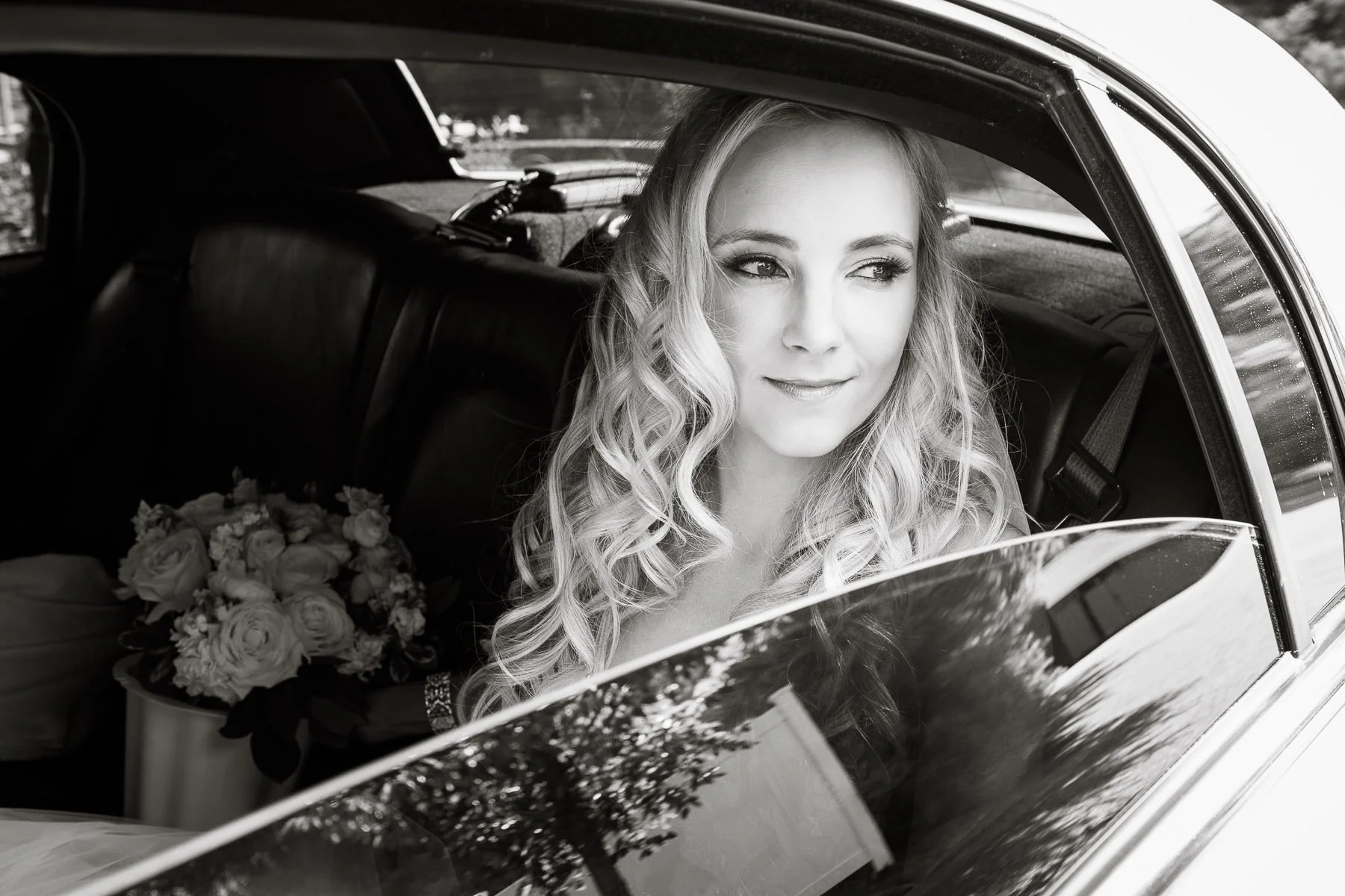 A bride sits in the back seat of a car and looks out the window before the ceremony, with her bouquet beside her. The black-and-white portrait captures a quiet, reflective wedding-day moment