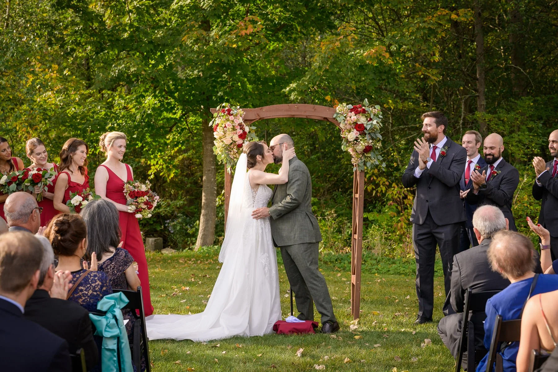 Chelsea and David share their first kiss during their outdoor wedding ceremony at the Inn at Manchester in Vermont. Guests and wedding party members applaud as the couple stands beneath a floral arbor on the lawn.