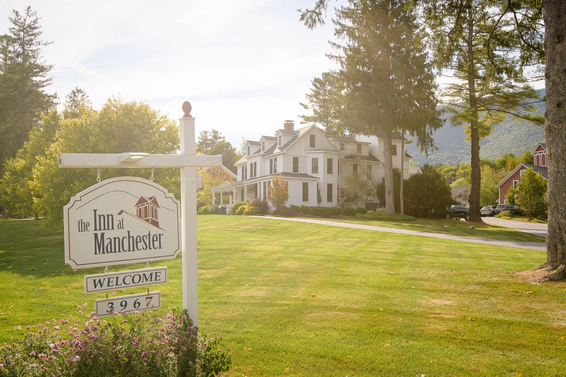 Exterior view of the Inn at Manchester in Vermont, with the welcome sign in the foreground and the white inn surrounded by lawns and trees. This Manchester wedding venue is shown in soft afternoon light with mountain views in the background.
