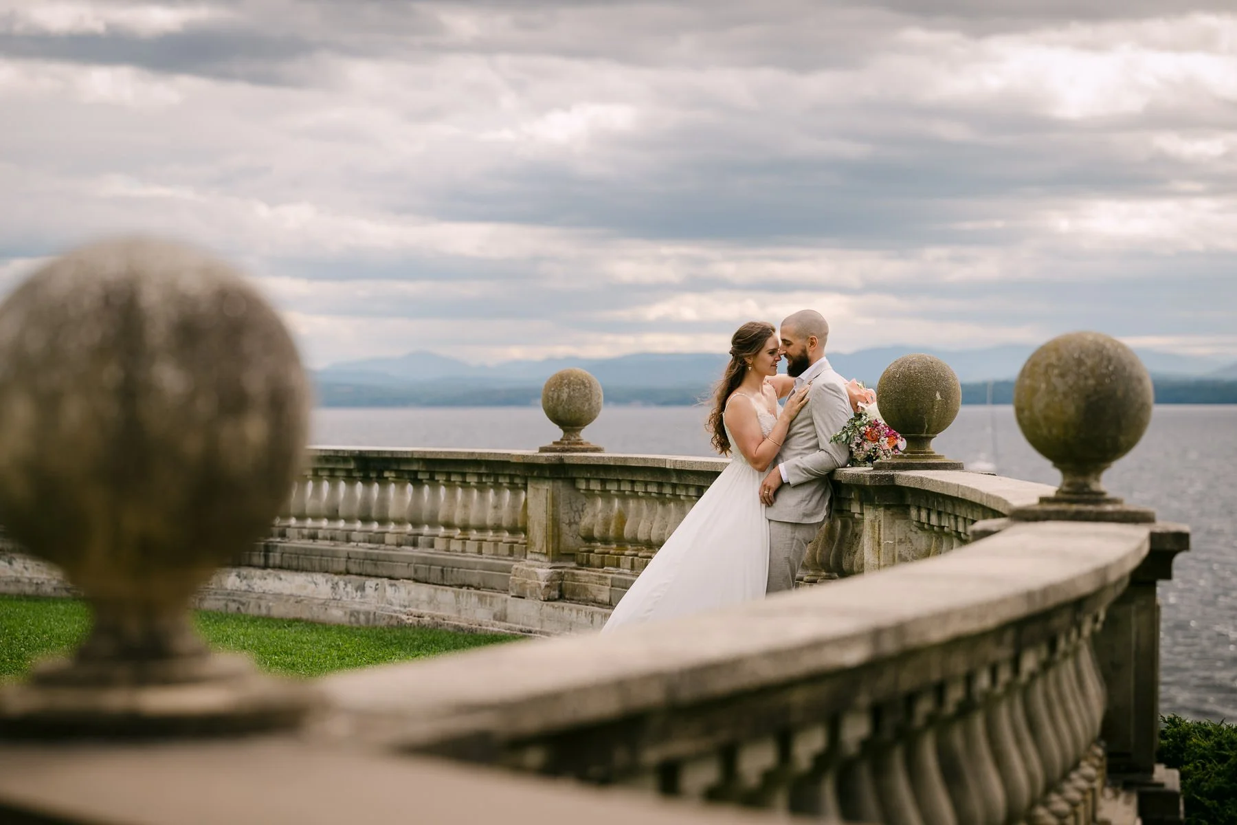 Bride and groom share a quiet portrait on the stone terrace at the Inn at Shelburne Farms in Shelburne, Vermont, with Lake Champlain and layered clouds in the background. A late-summer wedding portrait with a calm, scenic lakeside feel.