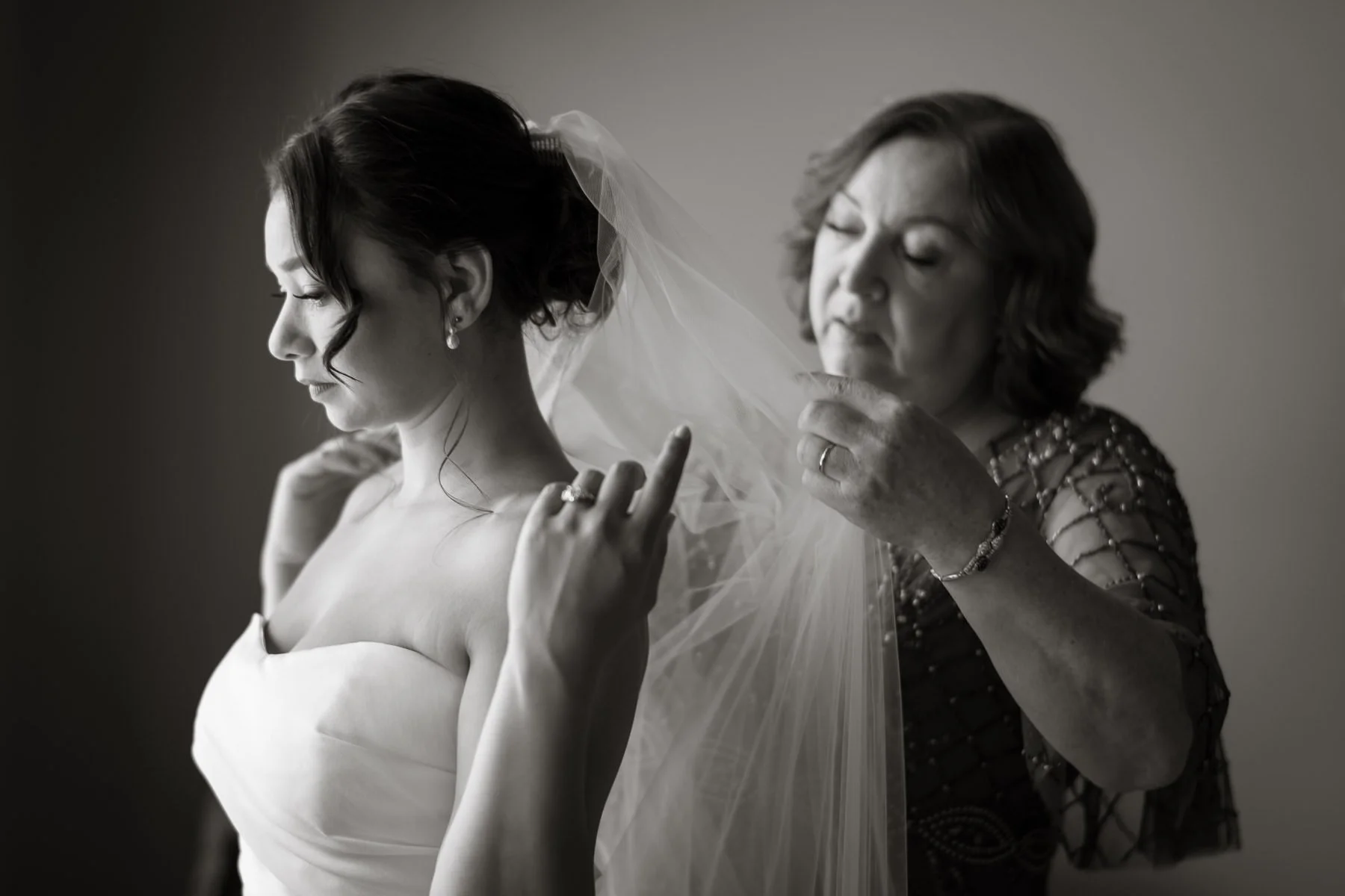 A bride stands by the window as an older woman helps adjust her veil during the getting-ready portion of the wedding day. The black-and-white image captures a quiet, intimate moment before the ceremony.