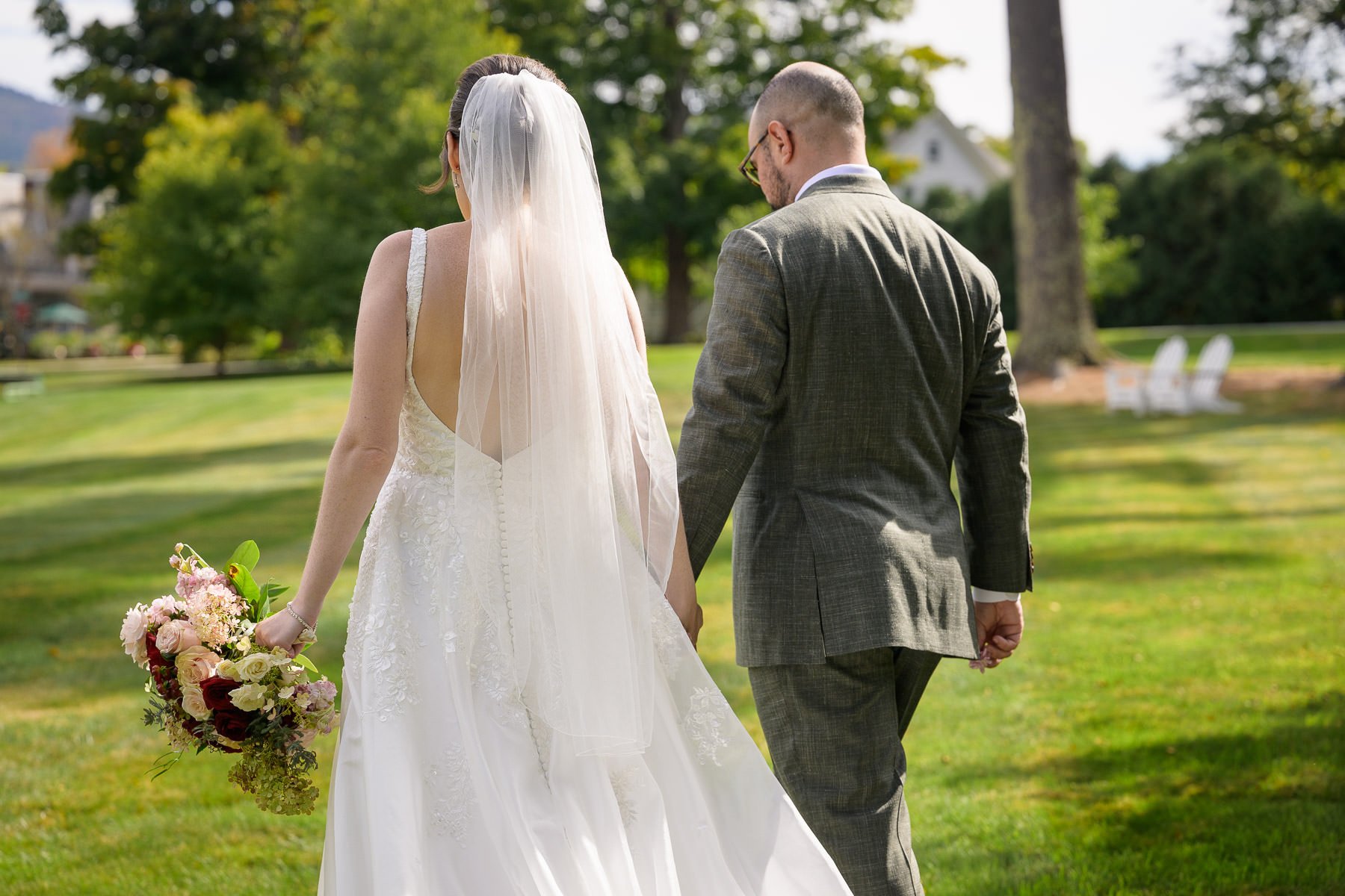 Chelsea and David walk hand in hand across the lawn at the Inn at Manchester in Vermont, seen from behind as Chelsea carries her bouquet and veil. The portrait captures a quiet just-married moment in the soft afternoon light.