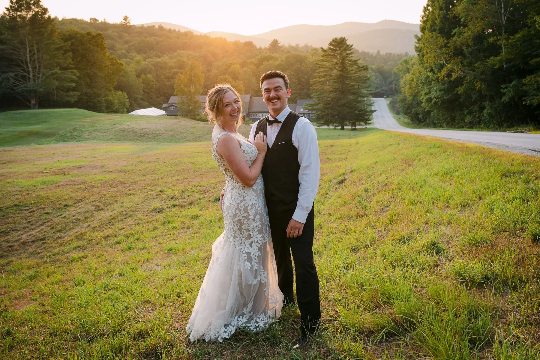 Lauren and Mike stand together in a grassy field at The Trailside Inn in Killington, Vermont, during sunset portraits with mountain views behind them. The warm early-August light gives the wedding portrait a relaxed, natural feel.