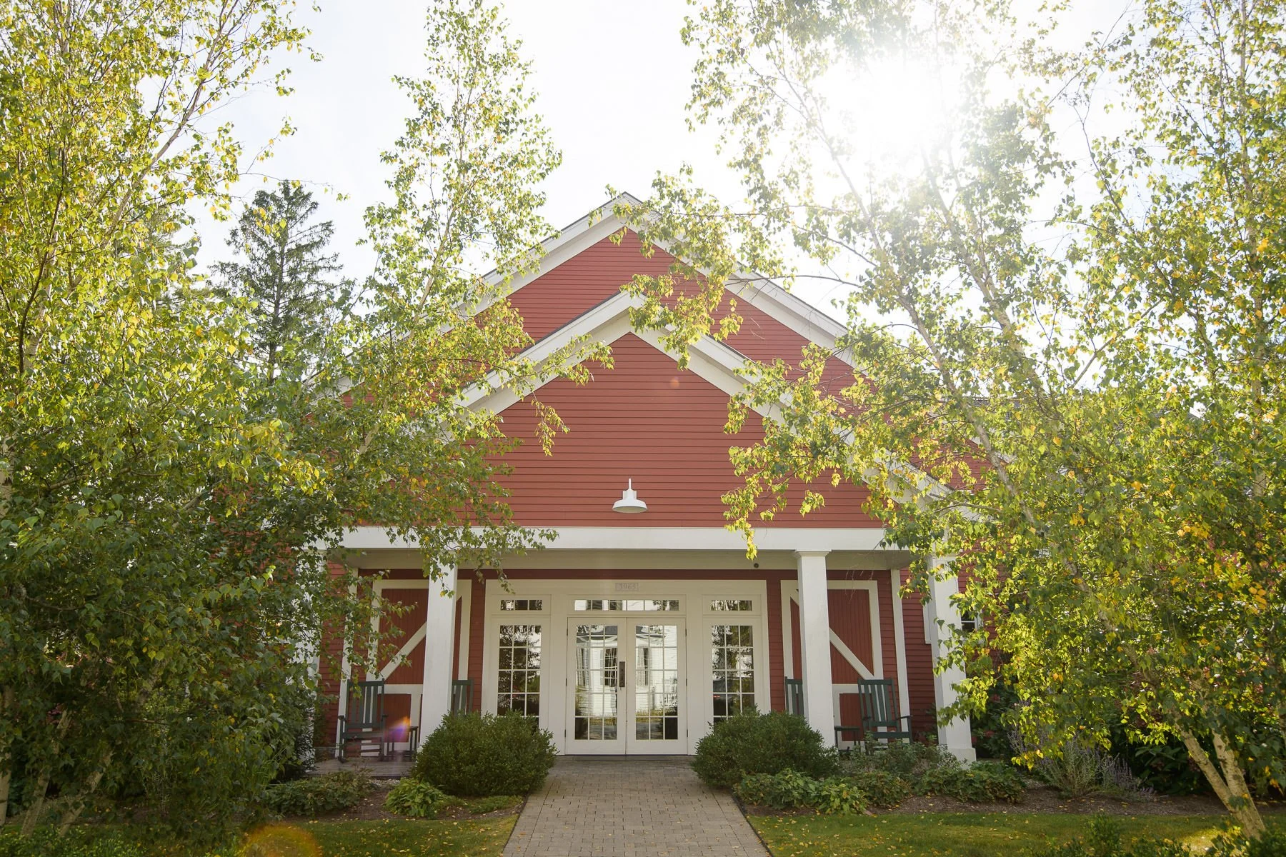 Exterior of the red barn at the Inn at Manchester in Vermont, framed by trees and bright afternoon light. The wedding reception space sits at the end of a brick walkway surrounded by greenery.