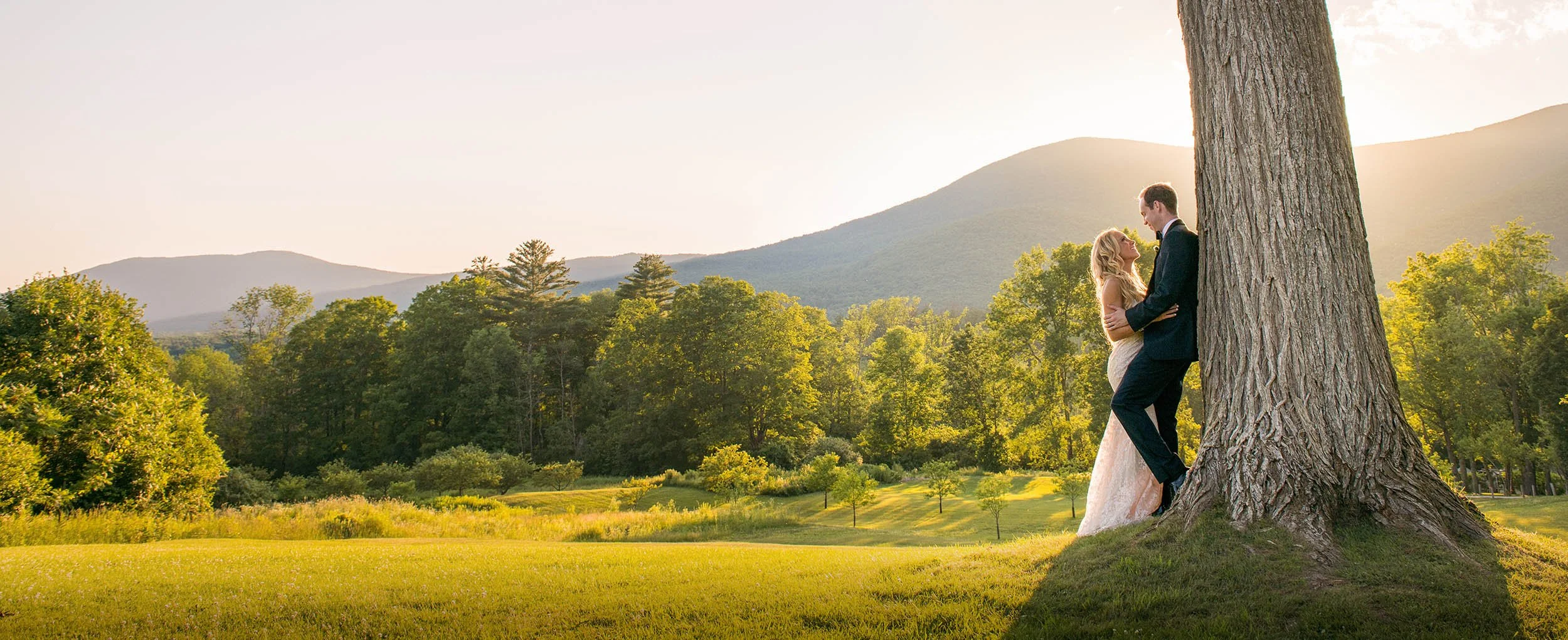 Bride and groom embrace beside a large tree at The Hildene in Vermont, with green hills and mountains glowing at sunset in early July.