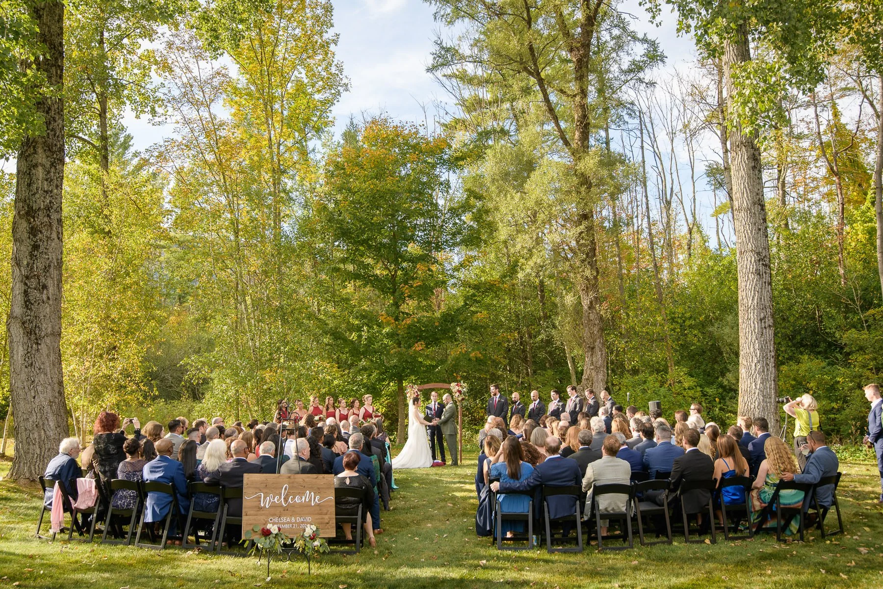 Chelsea and David’s outdoor wedding ceremony at the Inn at Manchester in Vermont, with guests seated on the lawn beneath tall trees as the couple stands at the altar. The wide view captures the natural setting and atmosphere of their ceremony.