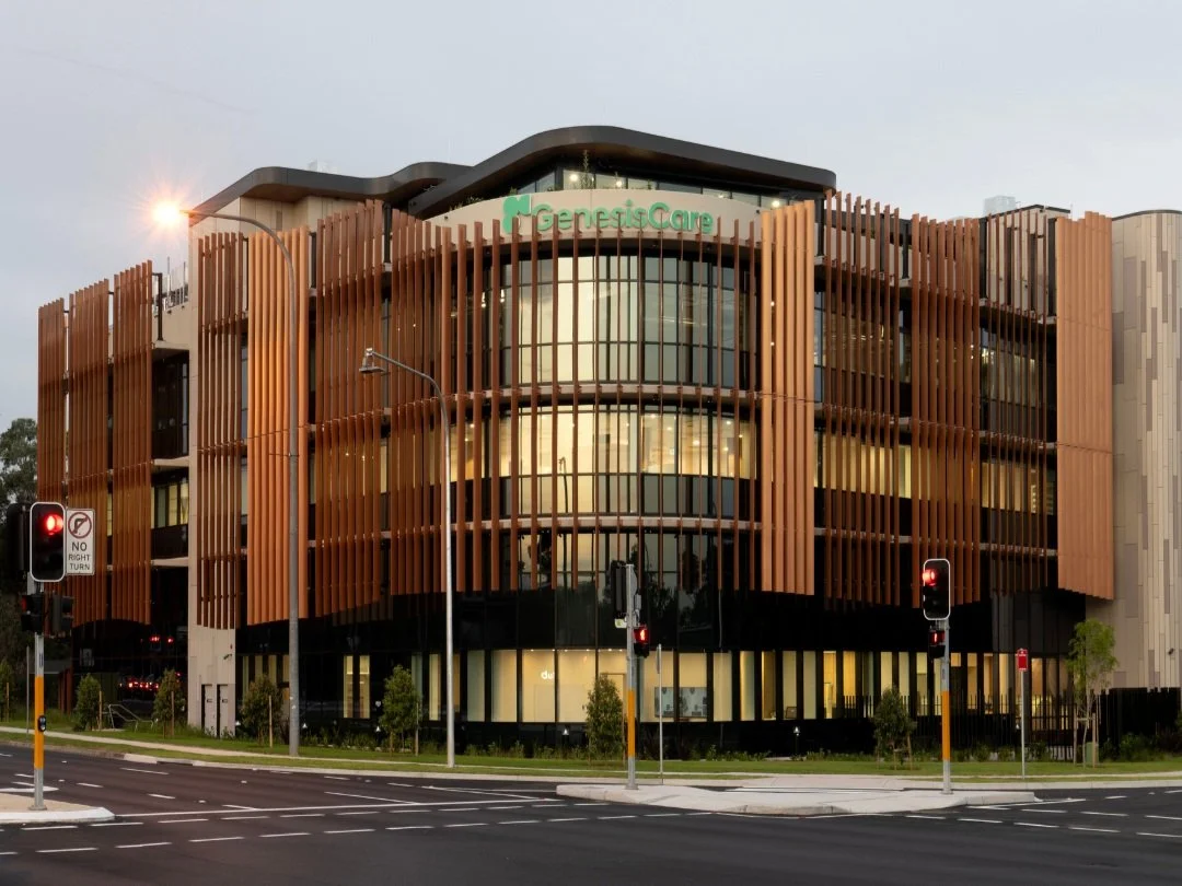 Modern medical office building with the sign 'Genetic Care' on top, featuring curved glass windows and vertical wooden slats exterior, situated at a city intersection with traffic lights and street signs.