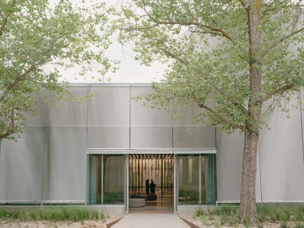 Modern building entrance framed by trees, with glass doors and a glimpse of the interior.